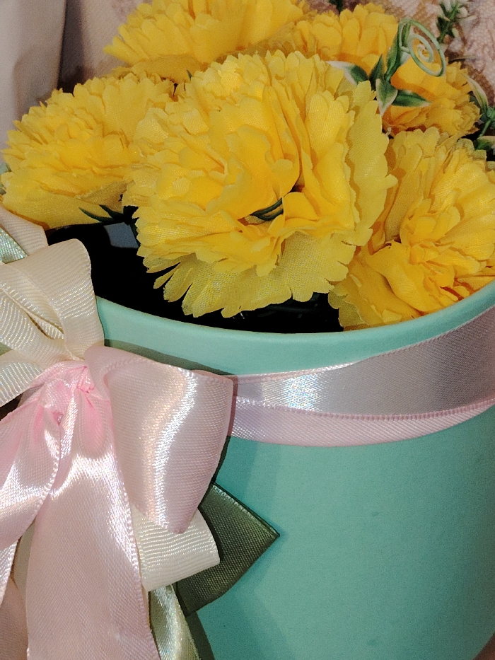 The image displays a close-up, high-angle view of a decorative floral arrangement situated in the city of Марковский, Russia. The arrangement consists of multiple artificial yellow flowers, characterized by dense, ruffled petals, resembling carnations. These flowers are contained within a light turquoise, cylindrical vessel with a smooth, matte finish. The container is embellished with multiple satin ribbons. A prominent bow, tied on the left side of the container, is composed of a light pink ribbon layered over a cream-colored ribbon. A separate, wider light pink satin ribbon encircles the upper circumference of the container. Segments of dark green satin ribbon are visible beneath the primary bow. No individuals or active interactions are present in the scene. The background is blurred, revealing indistinct light-toned surfaces, indicative of an indoor environment.