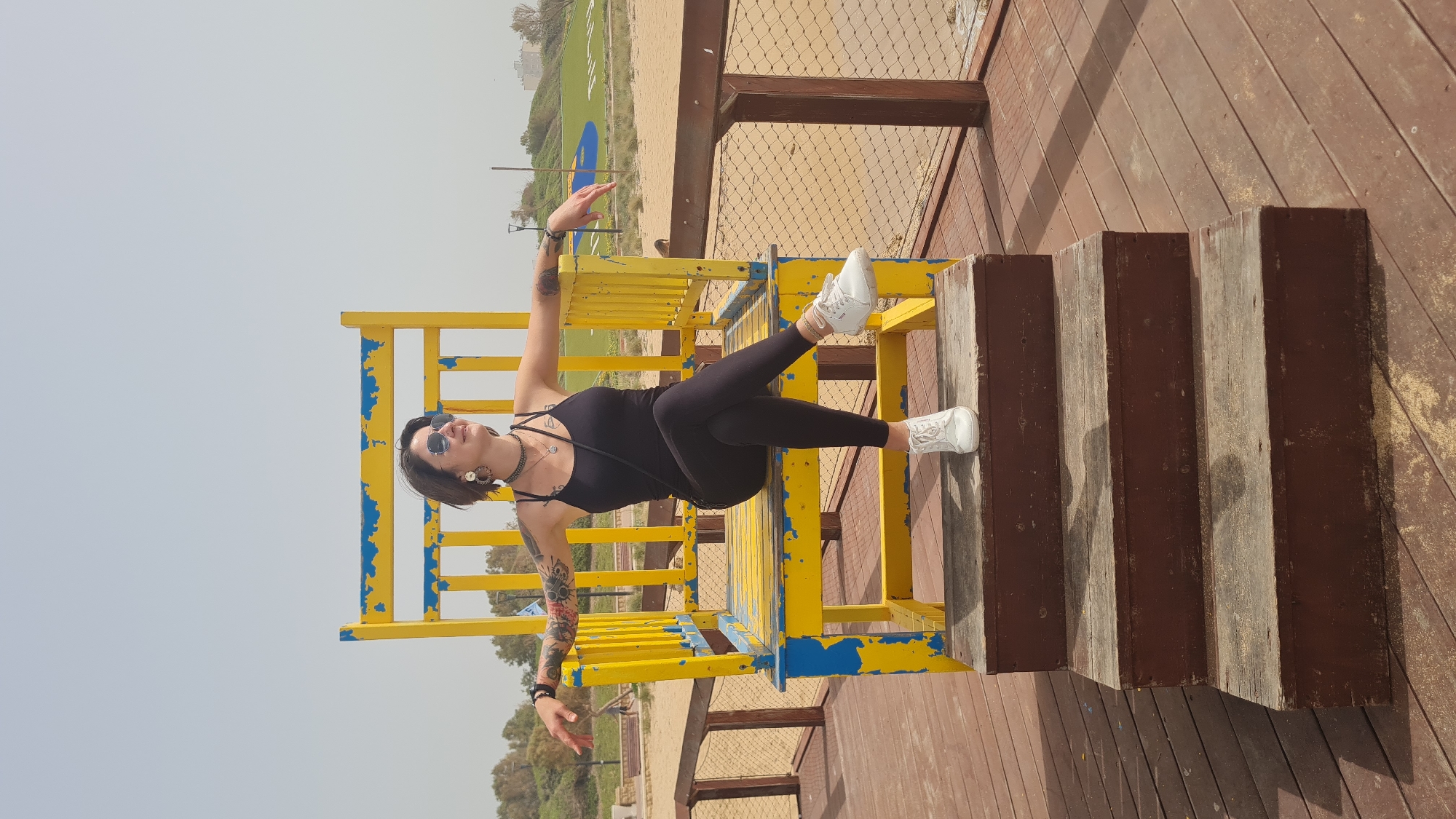 A female is positioned on an oversized yellow and blue chair. The chair is located on a wooden deck. The woman is wearing a black bodysuit, black leggings, and white sneakers. She is also wearing sunglasses and jewelry. She has short hair and visible tattoos on her arms. The background includes a sandy area with green grass, trees and buildings. The location is in the city of Unknown, Unknown.