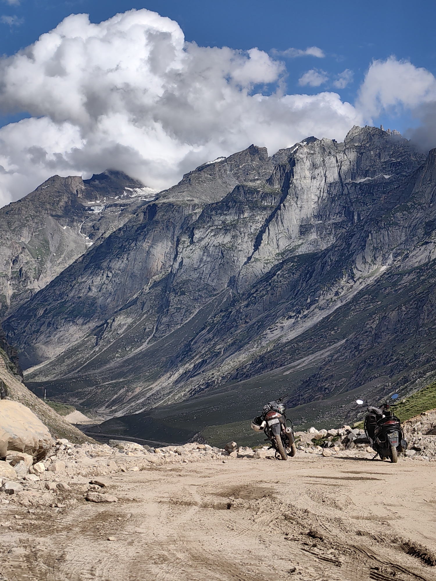 A dirt road winds through a rocky mountain landscape, leading towards a steep, rugged mountain range under a partly cloudy blue sky. Two motorcycles are parked on the side of the road. The motorcycle on the left is a dirt bike, parked on its kickstand, facing away from the camera. The motorcycle on the right is a scooter, parked on its kickstand, also facing away from the camera. The road is muddy and rutted in places. The mountains are covered in scree and sparse vegetation. The scene appears to be in a remote, mountainous area. The image is a vertical, full shot.