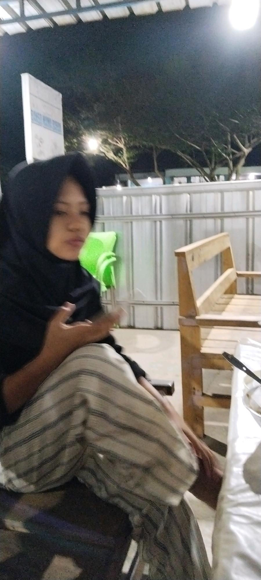 The image is a medium shot taken from a low angle at night, outdoors, in an unidentified location in Indonesia. A young woman wearing a black hijab and a striped skirt is seated on a wooden bench. She is looking down and to the left, with her mouth slightly ajar, in a pouting expression. Her left arm is bent, with her hand near her chest. To her right is a green plastic chair. Behind the woman, there is a corrugated metal fence. A wooden table with a white tablecloth is visible to the far right of the frame, with a glimpse of a serving utensil. In the background, there are trees with branches silhouetted against the night sky, and a white rectangular signpost. The overall lighting is dim, with artificial lights illuminating the scene from above.