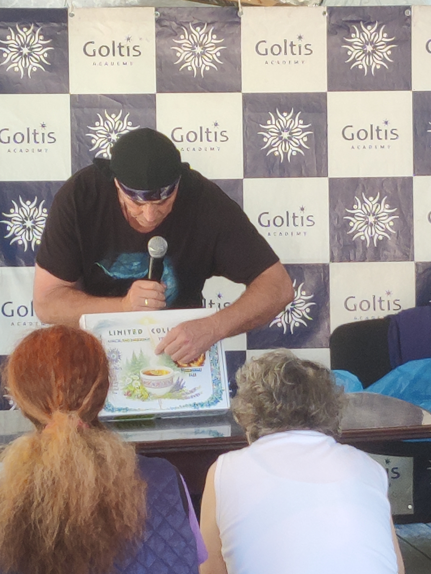 A man in a black t-shirt and black beanie is standing behind a table, holding a microphone in his right hand and pointing at a tray with a picture of a cup of tea. He has a ring on his right ring finger. In front of him, two women are seated, one with reddish-brown hair and the other with curly blonde hair wearing a white shirt. The background features a checkered banner with the text "Goltis Academy" repeated. The scene appears to be an indoor event, possibly a presentation or a signing. The image is taken from a slightly elevated angle, looking down at the subjects.