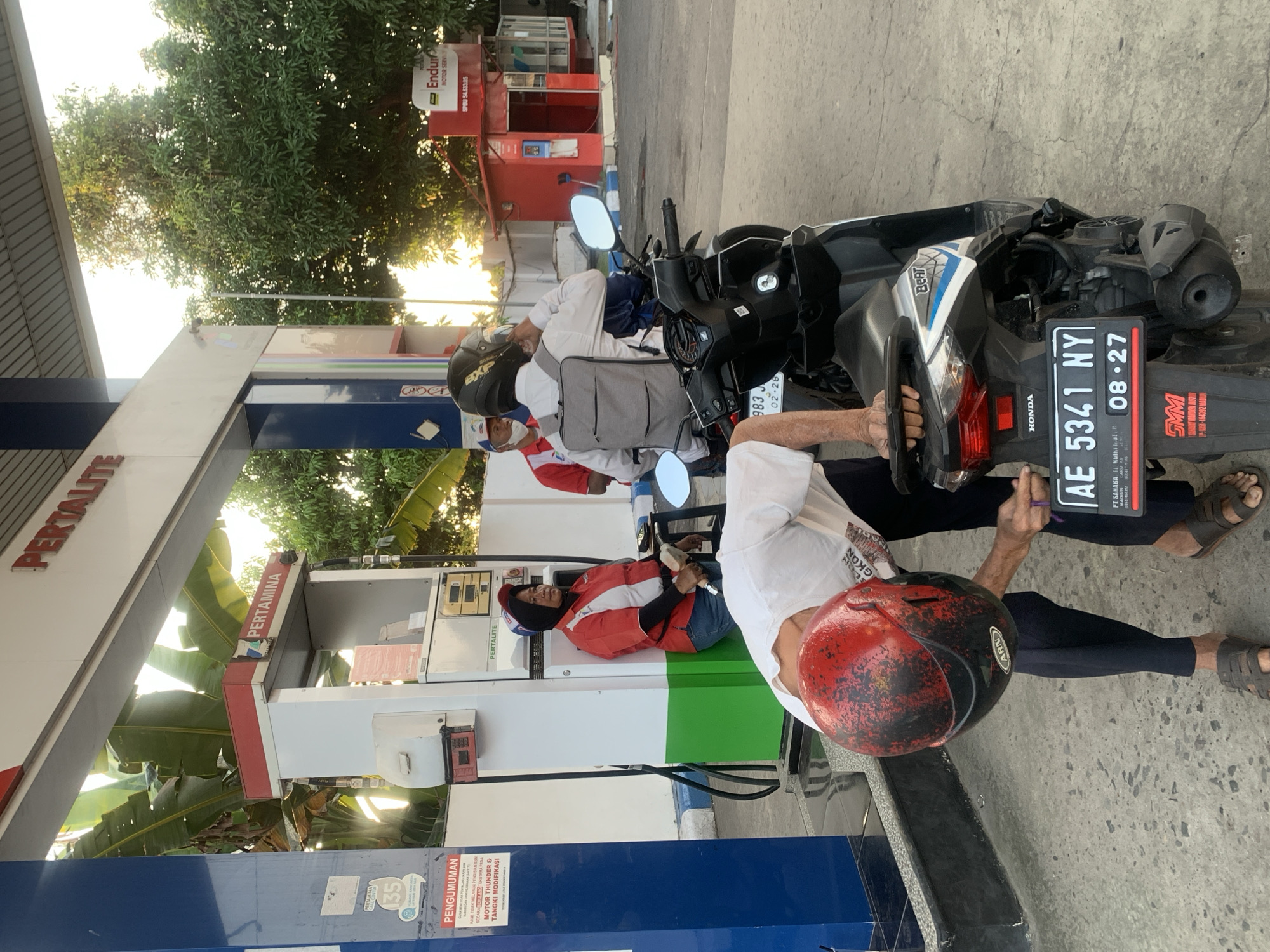 The image depicts a gas station in Mantren, Indonesia, illuminated by warm, low-angle sunlight, indicating either early morning or late afternoon.

In the foreground, an individual is bent over a black Honda Beat scooter. This person wears a white t-shirt, dark pants, and dark sandals, and holds a weathered red and black motorcycle helmet with "ARN" branding in their left hand. The scooter's license plate, "AE 5341 NY," is clearly visible, along with an expiration date "08-27" and a small "SMM" sticker on the rear fender.

To the left, a female gas station attendant stands beside a white and red fuel pump. She wears a red polo shirt with white accents, blue jeans, and a blue head covering (hijab), holding a fuel nozzle in her right hand. The fuel pump features the "PERTAMINA" logo and "PERTALITE" branding. An overhead canopy also displays "PERTALITE" in red lettering. A blue vertical column on the far left has a white sign reading "PENGUMUMAN" (Announcement) and partial text below indicating "MOTOR THUNDER & TANGKI MODIFIKASI."

In the midground, further to the right, a customer sits on a dark scooter, facing away from the camera. This individual wears a white long-sleeved shirt, a grey backpack, and a black full-face helmet with an "EXp" logo, appearing to adjust the helmet. Behind this customer, a male gas station attendant is partially visible, wearing a red polo shirt, a white undershirt, and a white face mask.

In the background on the right, a red kiosk-like building with a white sign reading "Enduro MOTOR SERVI" and "SPBU 54.633.35" is visible. Green trees with dense foliage are present in the background, along with large banana leaves visible behind the fuel pump on the left, suggesting a tropical environment. The ground is paved concrete, with a blue and white painted curb or barrier on the right side.