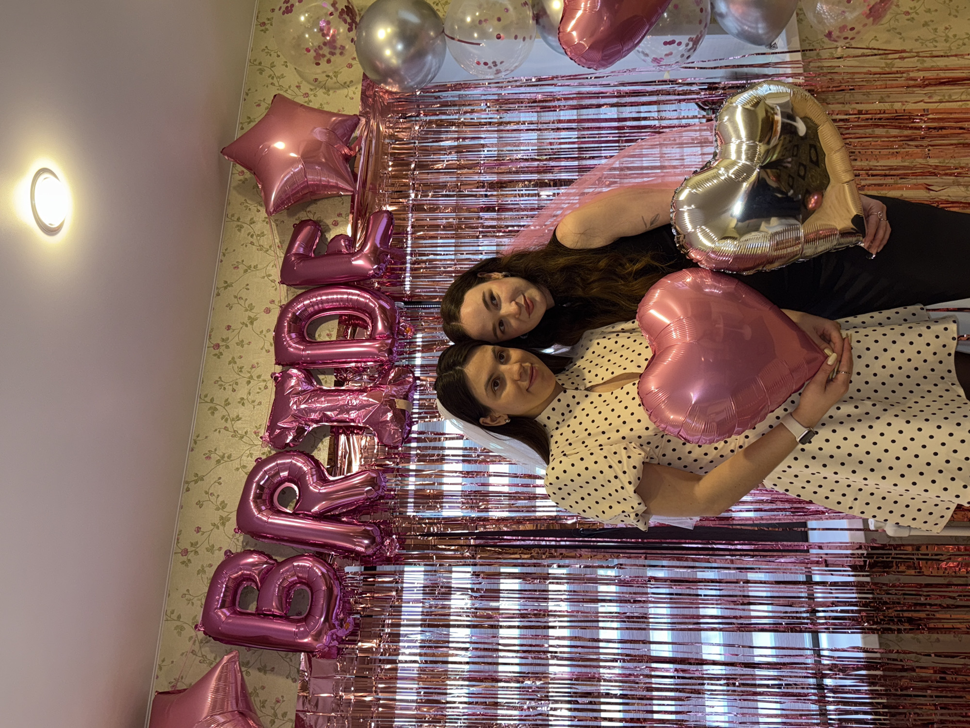Two young adult females are depicted in an indoor celebratory setting, identified by the prompt as located in Perm, Russia. Both individuals wear short veils, suggesting a bridal event. The female on the left wears a white dress with black polka dots, puffed sleeves, and a high collar, holding a large pink heart-shaped foil balloon with both hands. A white smartwatch is visible on her left wrist. The female on the right wears a black sleeveless garment and holds a large silver heart-shaped foil balloon with both hands.
The background features extensive decorations. Large pink foil letter balloons spelling "BRIDE" are centrally strung across a pink shimmering tinsel curtain, which covers the majority of the wall. Pink star-shaped foil balloons are positioned to the left and right of the "BRIDE" letters. To the far right, a stack of various balloons includes clear latex balloons filled with pink confetti, spherical silver foil balloons, and additional pink heart-shaped foil balloons. Behind the tinsel curtain on the left, horizontal window blinds are partially visible. The wall above the tinsel curtain is covered with light-colored wallpaper featuring a subtle floral or vine pattern. The ceiling is white, with a single recessed light fixture emitting a warm glow in the upper central portion of the frame.