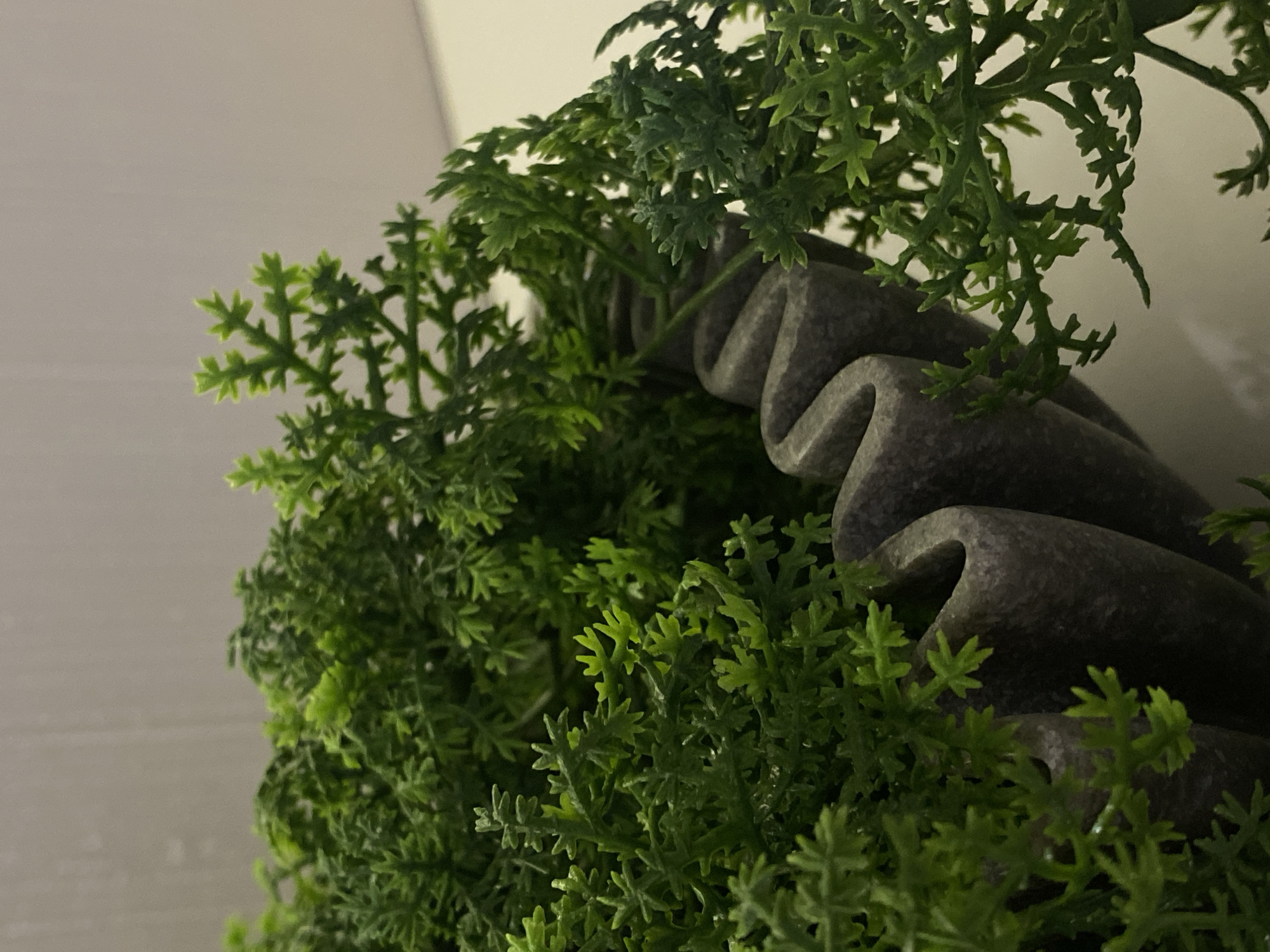 The scene, situated in the city of Votkinsk, Russia, depicts a close-up of a dense arrangement of artificial green foliage within a distinctive container. The foliage consists of numerous small, intricately branched, bright green leaves resembling a fern or conifer, with some areas appearing darker green due to shadowing. The plant material is synthetic. The container holding or bordering the foliage is dark gray, featuring a pronounced, uniform wavy or corrugated texture along its visible edge, creating deep, vertical undulations. The material appears non-reflective, possibly stone or concrete. The background is a plain, light-colored wall or surface, suggesting an indoor environment. No individuals or other subjects are present. The lighting is even, with illumination from the upper right.
