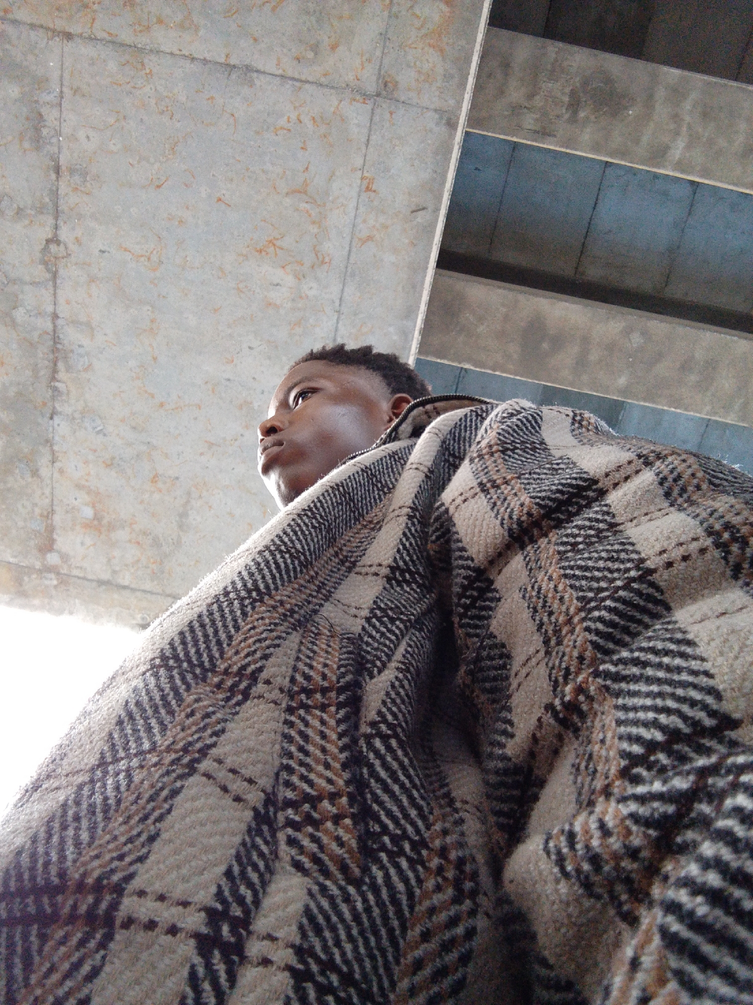 A low-angle medium shot shows a dark-skinned young man looking up and to the left, wearing a brown and black plaid jacket. The man has short, dark hair. The jacket is covering most of his body. Above him is a concrete ceiling with some staining. In the background is a section of concrete wall with blue paint. The picture may be taken in an underpass or other open infrastructure, possibly in the city of Lekki, Nigeria.
