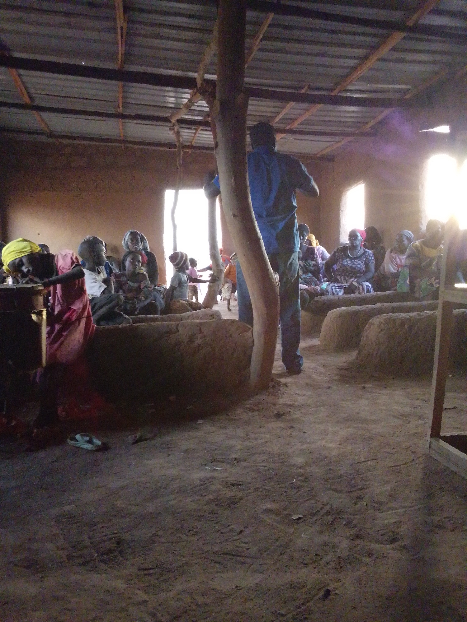 The image shows the interior of a rudimentary building in Mulke Barikin Sayejo, Nigeria. The structure has mud walls, a corrugated metal roof supported by wooden beams, and several window openings. Inside, a group of people, predominantly women and children, are seated on raised earthen platforms arranged in rows. A man in a blue shirt stands, partly obscured by a wooden post, facing the group. A variety of clothing is visible, and a pair of sandals lies on the dirt floor in the foreground. The overall lighting is dim, with strong sunlight streaming through the windows.