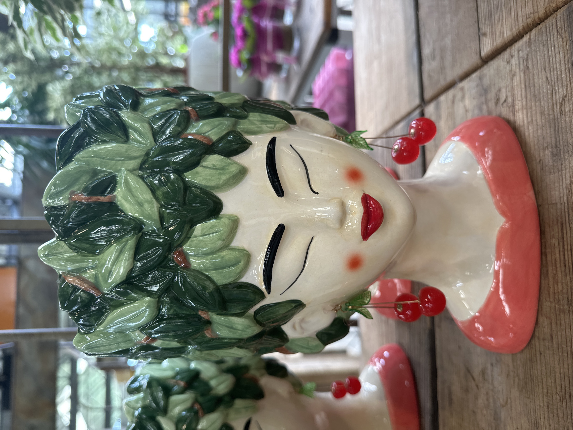 A close-up, eye-level shot shows a ceramic planter shaped like a woman's head. The planter has a cream-colored face with closed black eyes, rosy cheeks, and red lips. A crown of green leaves adorns her head. Dangling from her ears are earrings resembling cherries. The planter is positioned on a wooden surface. In the background, another similar planter is visible to the left, and blurred pink flowers and greenery are seen further back. The scene appears to be indoors, possibly in a shop or greenhouse.