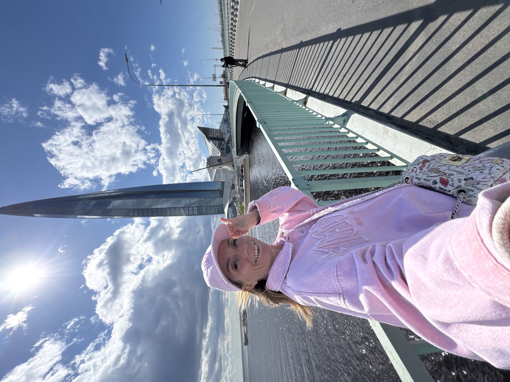 A woman dressed in a pink outfit, including a matching hat, poses on a bridge with a view of a futuristic skyscraper. The skyscraper is the Lakhta Center in Saint Petersburg, Russia. The woman is smiling, wearing large hoop earrings, and making a peace sign with her fingers. A patterned purse rests on the bridge railing beside her. The bridge is lined with a green metal railing, casting long shadows on the asphalt path. In the distance, a lone figure walks along the bridge. The sky is filled with puffy white clouds against a bright blue backdrop. The sun shines brightly, creating a starburst effect. Below the bridge is a body of water.
