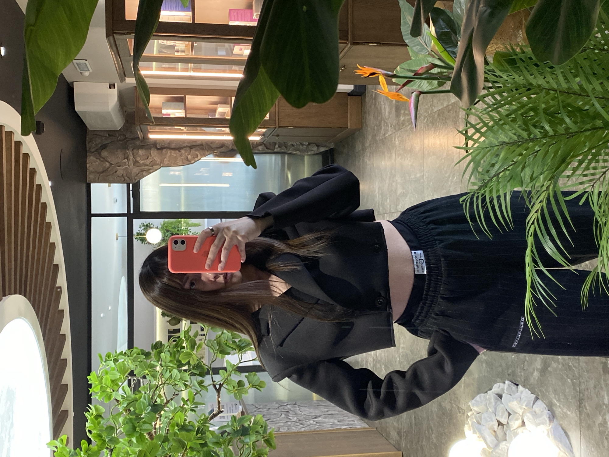 A young person with long brown hair is captured in a mirror selfie, standing in a modern indoor space in Votkinsk, Russia. The individual wears a black cropped blazer over a black top, revealing a bare midriff, and loose-fitting black pinstriped pants with an elastic waistband. A bright coral-orange smartphone, held in their right hand, partially obscures their face as they take the photo. A ring is visible on their left index finger.
The setting appears to be a stylish commercial or public establishment, possibly a cafe or retail space, characterized by lush greenery and contemporary design elements. Large, tropical-leafed plants, fern-like foliage, and a plant with small, round green leaves are strategically placed throughout the room, including a vibrant orange and purple Bird of Paradise flower. The floor is a light-colored, textured surface. Architectural details include a striking arched structure with vertical wooden slats on the left, a section of wall clad in rough, light-colored stone, and glass display cases or shelves in the background. Overhead, various lighting fixtures, including strip lights, illuminate the space. In the foreground on the right, there is a decorative arrangement of white, angular rocks with small embedded lights.
Visible text on the person's clothing includes a white tag on the elastic waistband of their pants that reads "ORIGINAL" with additional, smaller text below it. Further down the left pant leg, there is white text, partially obscured, which appears to say "METEROCLO M" or similar. The overall ambiance is bright and modern, suggesting a daytime interior shot.