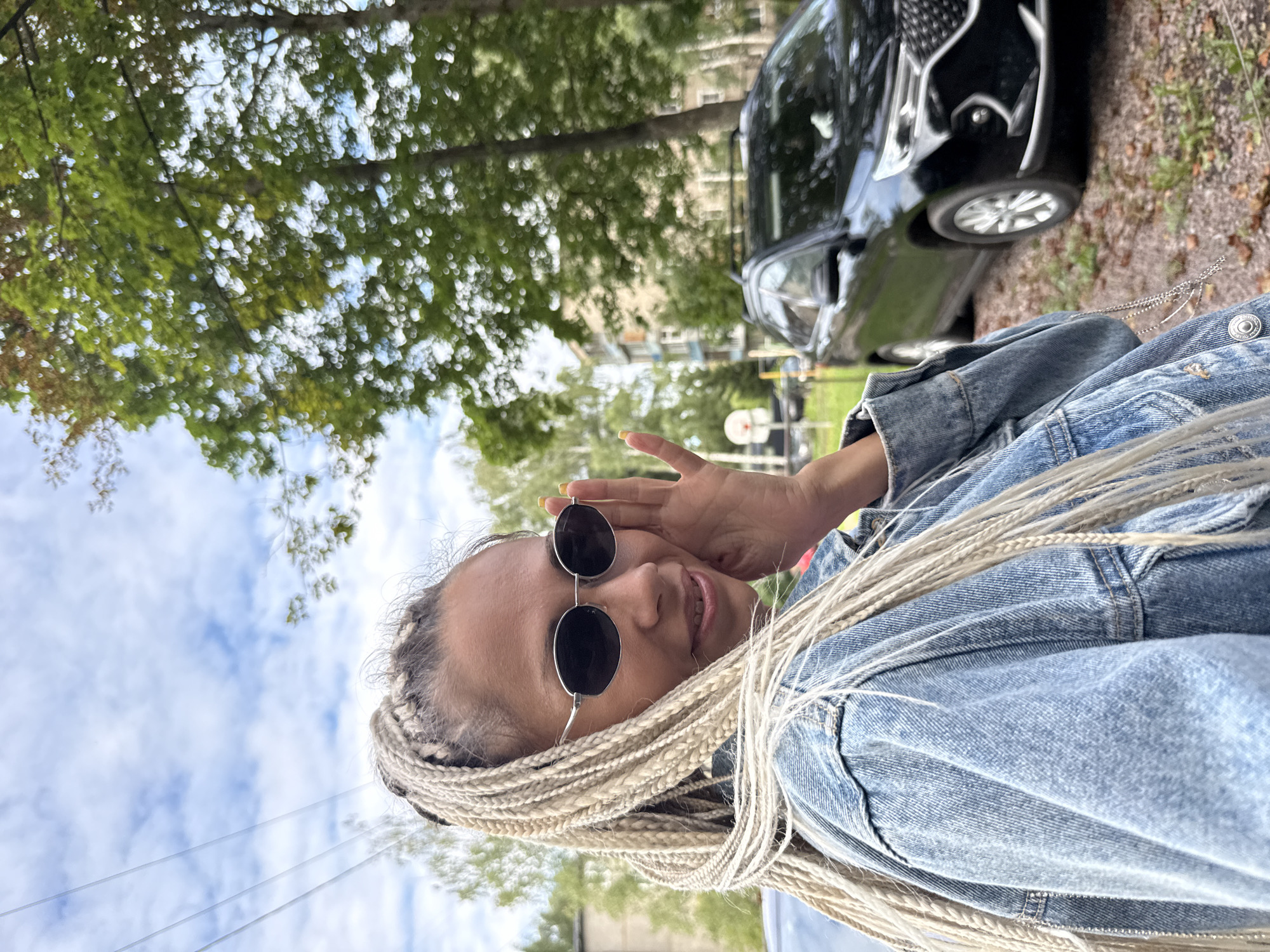In Rusanovo, Russia, a woman with long blonde braids and sunglasses is captured in a medium shot, leaning her head towards her right shoulder and touching her sunglasses with her left hand. She is wearing a denim jacket, and her blonde hair is styled in numerous braids that cascade down her shoulders and back. The background reveals a street scene with a black SUV parked on the right side, a building in the distance, and trees with green foliage. The sky above is partly cloudy, with visible power lines. The overall setting appears to be an outdoor urban environment on a bright day.