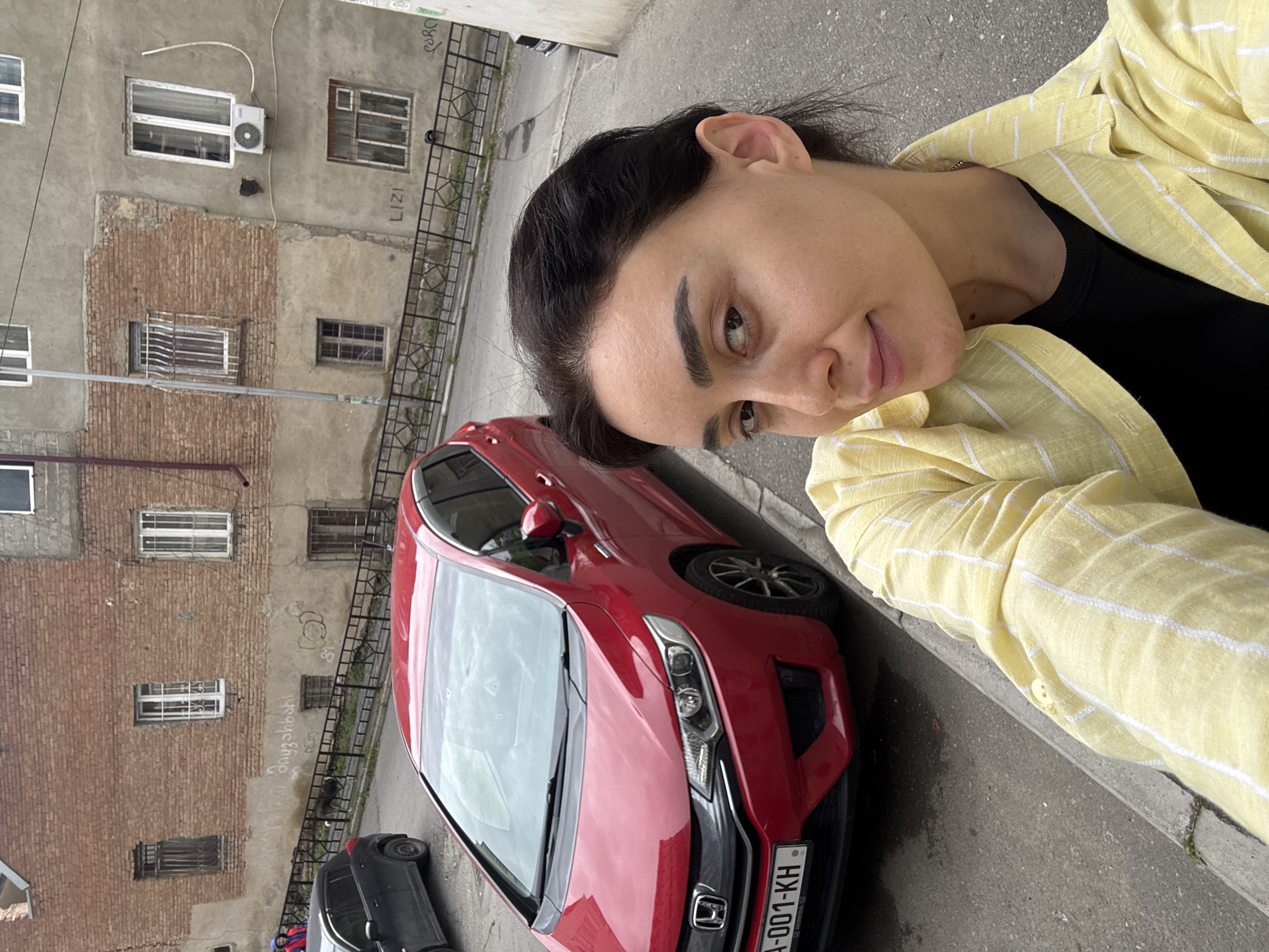 This outdoor selfie-style image features a young woman in the foreground, captured in an urban setting during what appears to be an overcast daytime. The woman has dark hair and a neutral to slightly smiling expression, looking directly at the camera. She is wearing a black top under a light yellow button-up shirt with white vertical stripes.
Behind her, a red Honda Jazz (or Fit) is parked on the street. Its Georgian license plate, "A-001-KH", is clearly visible. A small section of another dark-colored vehicle is also visible further down the street.
The background is dominated by an older, multi-story building constructed with a mix of exposed red brick and patched-up concrete/stucco walls. Several windows with varying designs, some featuring metal grates, are scattered across the facade. An outdoor air conditioning unit is mounted on the upper part of the building. A black metal fence or railing runs along the street in front of the building, which is paved with asphalt and has a concrete curb.
Visible text includes the car's license plate "A-001-KH". Graffiti on the building includes the Latin script word "LIZI", "PORQ", and other words in Georgian script that are less distinct. The overall scene provides a glimpse into a typical streetscape in Tbilisi, Georgia, as suggested by the car's license plate and architectural style.