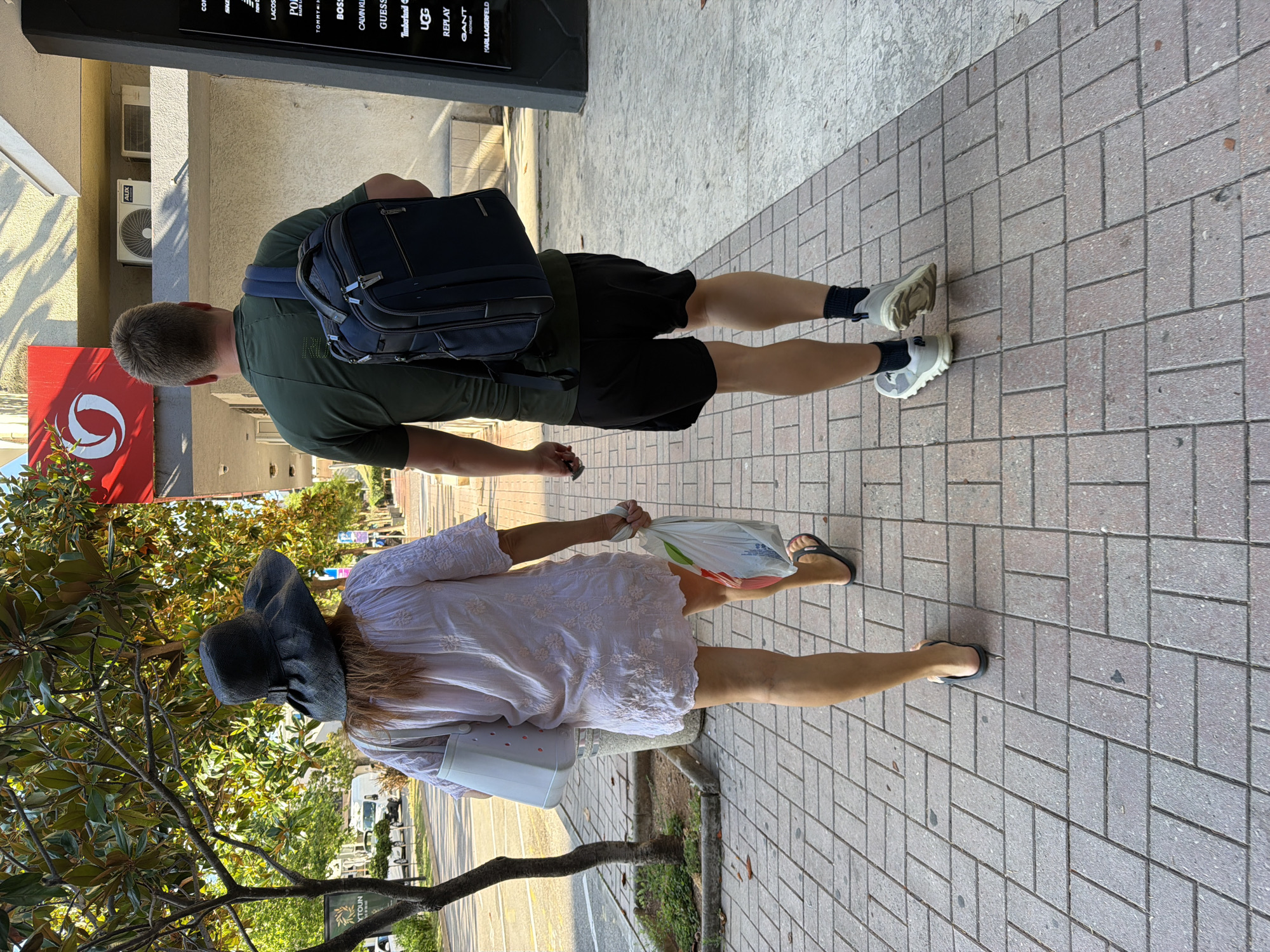 Two individuals, a woman and a man, are walking away from the camera on a paved pedestrian walkway in Budva, Montenegro. The woman, on the left, wears a wide-brimmed dark hat, a light-colored, short-sleeved, embroidered dress, and black flip-flops. She carries a white bag on her left shoulder and a plastic grocery bag in her right hand. The man, on the right, is wearing a dark green t-shirt, black shorts, and white athletic shoes with black socks. He has a dark blue backpack on his back. They are walking past shops. A red sign with a white circular logo is visible on the building behind them, and a black directory board with brand names is on the right. Trees with green foliage line the left side of the walkway, with a street and parked vehicles visible in the background. The sun casts shadows on the walkway and buildings.
