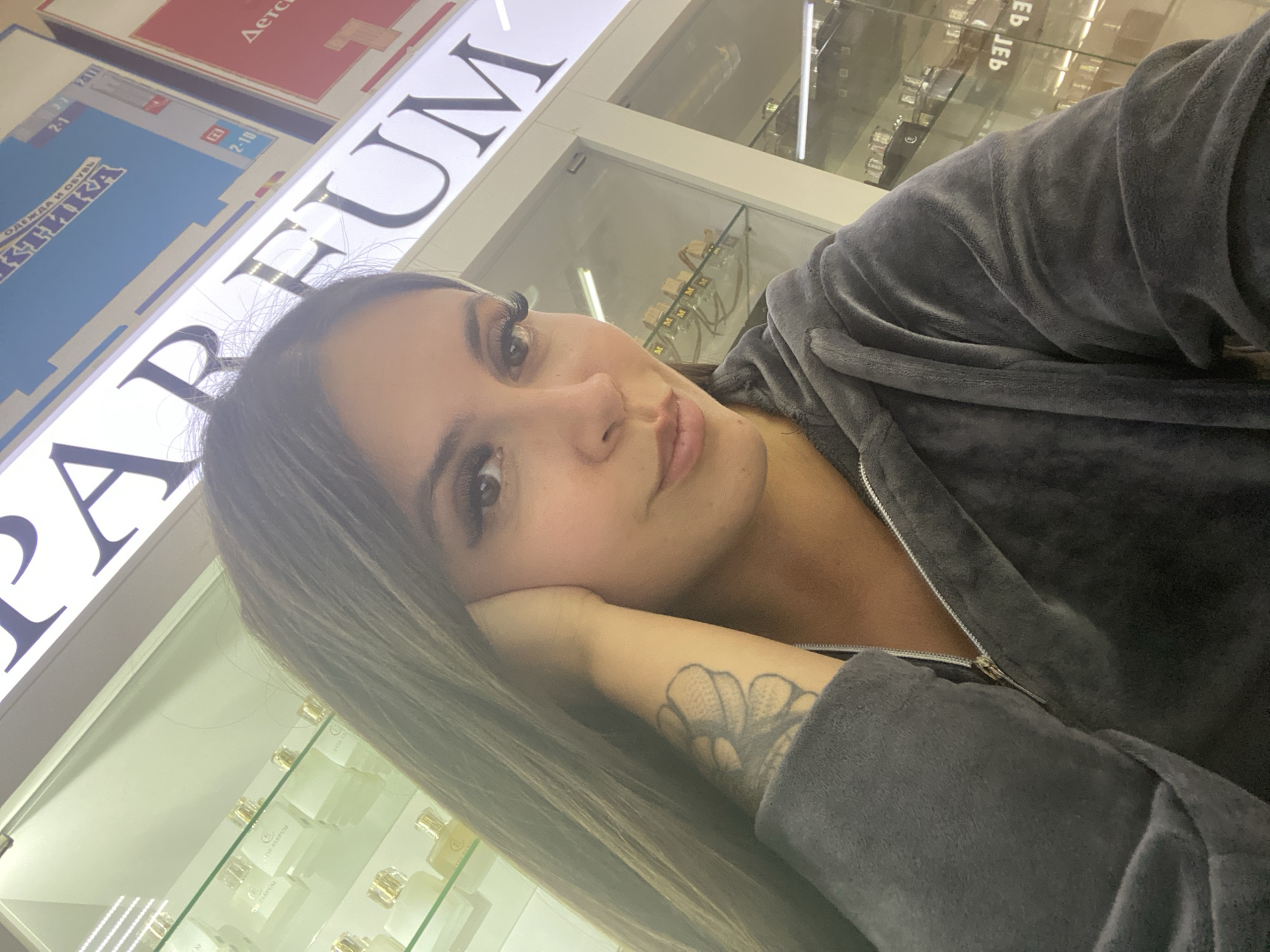 A medium shot photograph captures a woman with long, dark hair and dramatic makeup, including long, false eyelashes. She is resting her left hand on her cheek and her left arm is visible, showing a tattoo of a flower on her forearm. She is wearing a grey, zippered hoodie.
The background suggests a retail environment, possibly a department store or a duty-free shop. A large illuminated sign that partially reads "PARFUM" is visible behind her. Shelves displaying numerous perfume bottles are also present. In the far background, further signs and reflections in glass surfaces can be observed. The lighting is bright, typical of indoor retail spaces.
The provided context that this image is located in the city of Chaikovsky, Russia, is not visually verifiable from the image itself. There are no recognizable landmarks or signage that specifically identify the location as Chaikovsky.