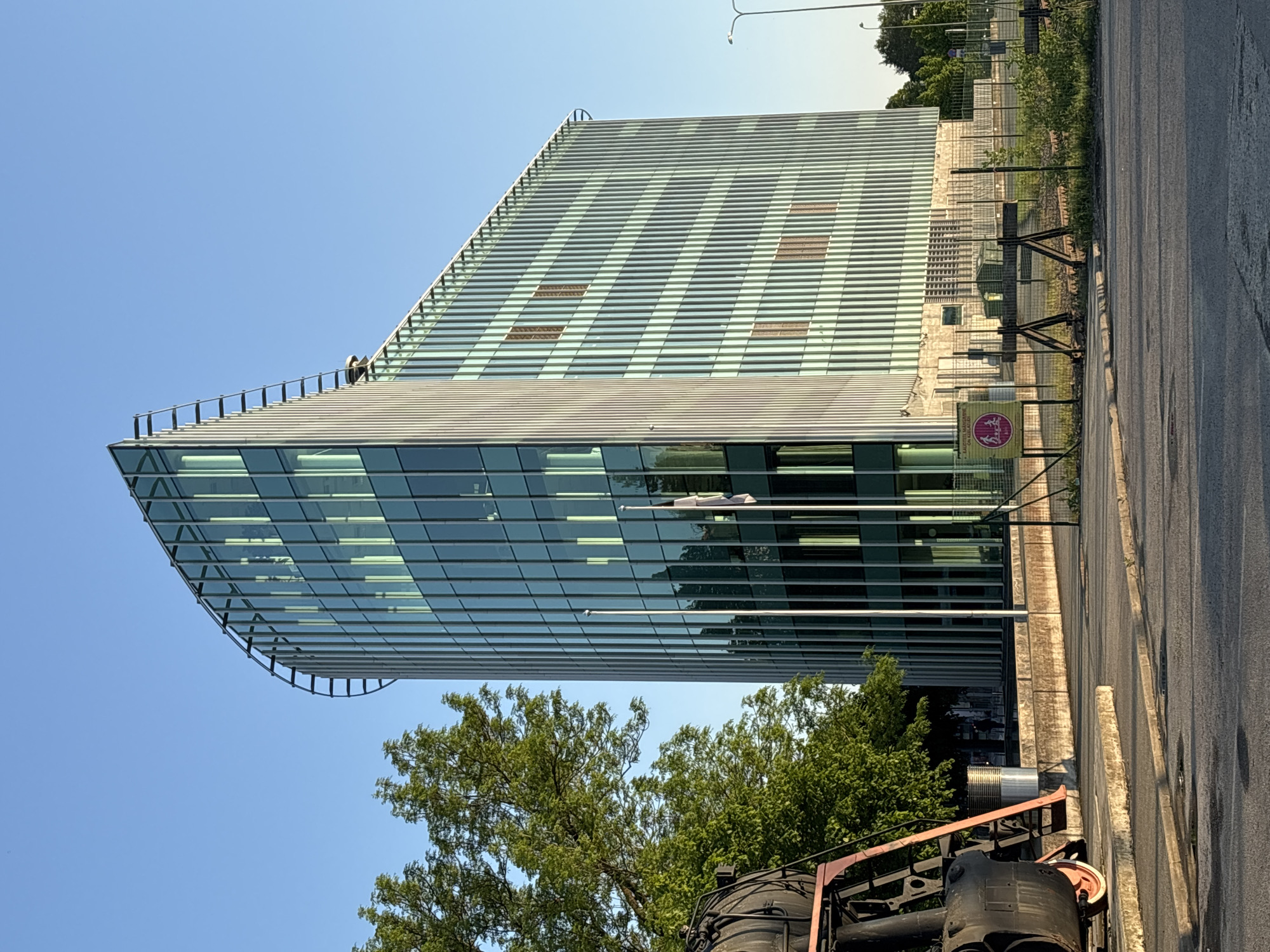 The image displays a modern, multi-story building with a distinctive architectural design, featuring extensive glass facades and horizontal louvers that provide sun shading. The building has a prominent angular corner, and a metal railing outlines its roofline. Two small figures, possibly maintenance workers, are visible on the roof near this corner, against a clear blue sky.
In the foreground, partially visible on the bottom right, is the front section of an old, dark-colored steam locomotive. An asphalt road runs along the right side of the image, separated from the building's immediate vicinity by a chain-link fence and some green foliage.
Attached to the fence is a yellow warning sign. The visible text on the sign includes "Hoiatus! Liikumine raudteel keelatud! Trahvi ähvardusel." in Estonian, and "Warning! Movement on railway forbidden! Under penalty of fine." in English, with additional Russian text. This indicates the presence of a railway line and suggests the setting is likely a museum or a historical park in Tallinn, Estonia. The scene is bathed in bright daylight under a clear sky, suggesting good weather conditions during the day. Lush green trees are visible in the lower part of the frame, contrasting with the modern architecture and industrial elements.