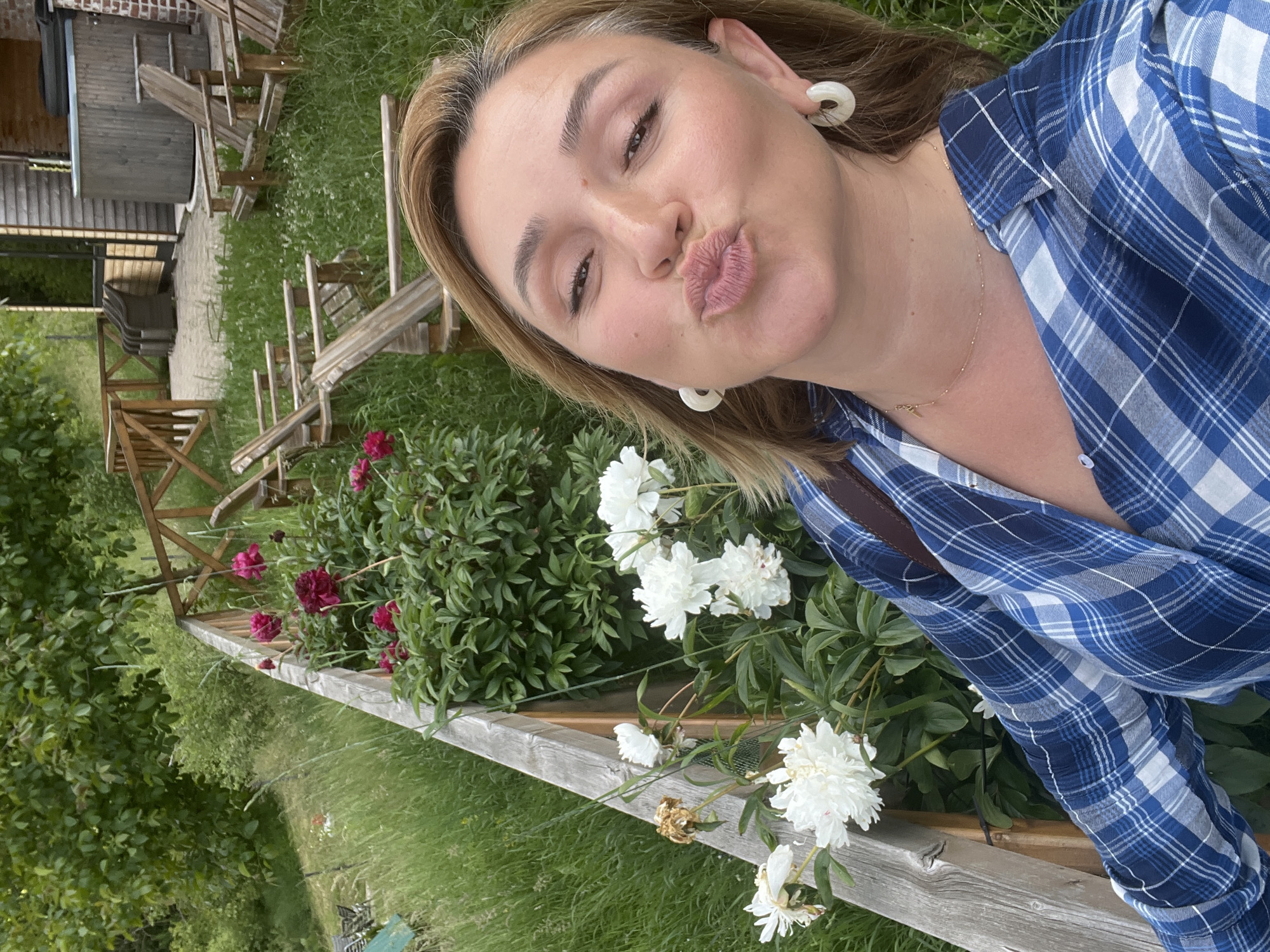 A woman is taking a selfie in a grassy outdoor area with a wooden fence and a building in the background. The woman has blonde hair and is wearing a blue and white plaid shirt and large white hoop earrings. She is puckering her lips for the camera. Behind her, there are white and pink flowers, a wooden fence, and several wooden chairs. A wooden building with a hot tub is visible in the background.