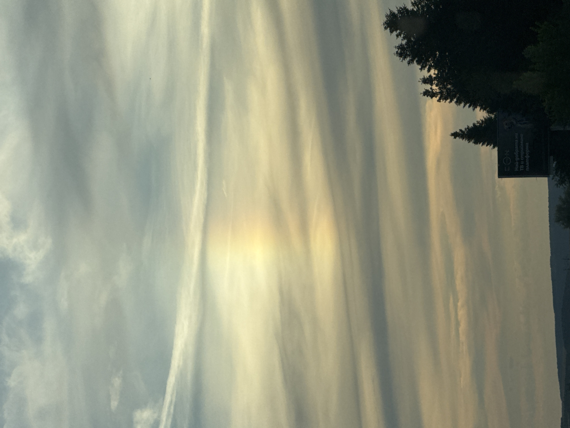 The image shows a natural phenomenon of a sun pillar, which appears as a vertical shaft of light extending upwards from the sun. The sun pillar is colored in pastel shades of yellow, orange, and pink, with a rainbow effect. It is visible in the sky above Gabarnitsa, Bulgaria, over a landscape of rolling hills and trees. The sky is filled with wispy clouds, and the overall atmosphere is serene and peaceful. There are no people or specific objects in the immediate vicinity of the sun pillar, but in the lower right corner of the image, there is a billboard with some text in Cyrillic script, and a silhouette of trees.