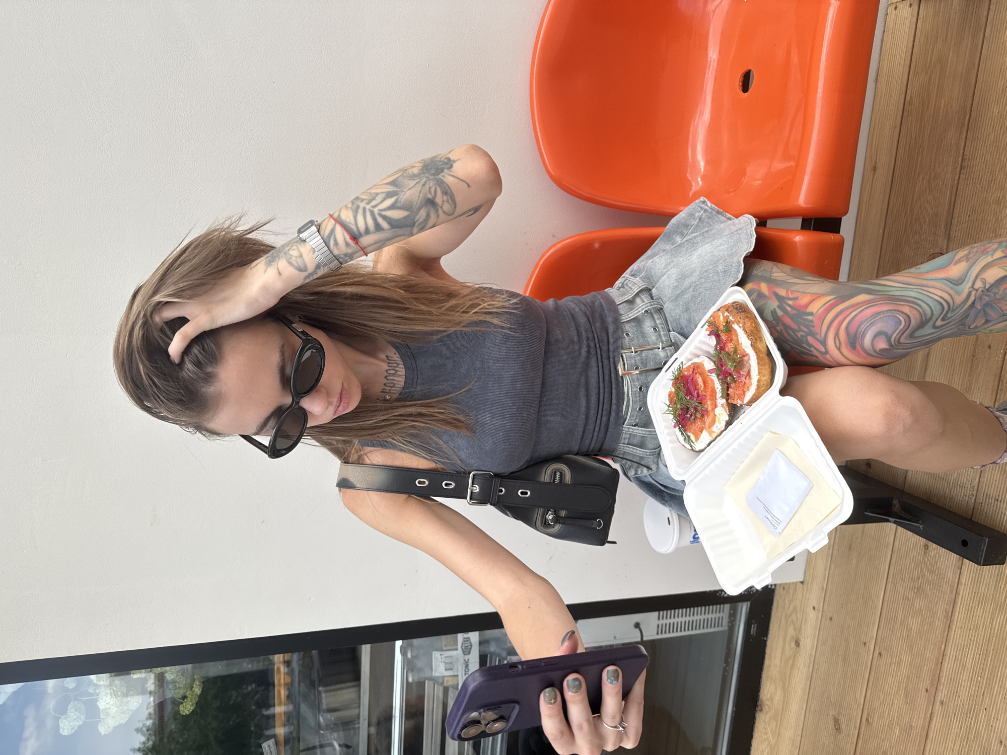 A woman is seated on an orange chair outside a building in Moscow, Russia. She is wearing a gray tank top and denim shorts, and has extensive tattoos on her arms and legs. She is holding a smartphone in her left hand and running her right hand through her hair. A black purse is slung over her left shoulder. A white takeout container holding two open-faced sandwiches with salmon and dill is resting on her lap. A small white cup is also on her lap, next to the food container. The background shows a white wall and a wooden deck. A window with a reflection of greenery is visible near her hand holding the phone.