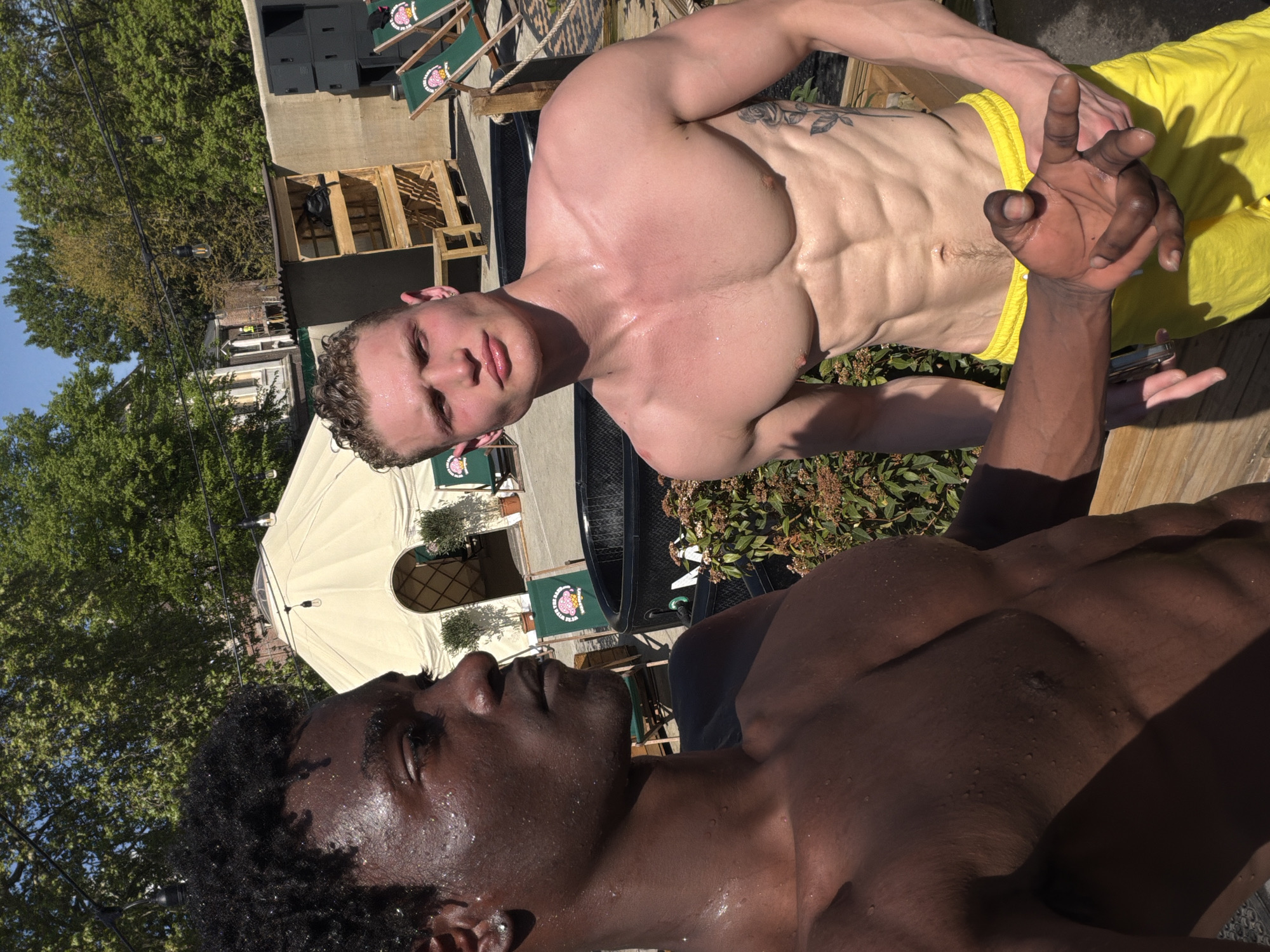 This outdoor portrait features two shirtless men in what appears to be a sunny urban setting in London, United Kingdom, possibly a rooftop or terrace. Both men are visibly wet, with water droplets on their skin, suggesting recent exposure to water or intense heat/activity.
In the foreground and to the right, a man with a darker skin tone, short curly black hair, and an athletic build is partially visible, primarily his head, shoulders, and chest. He is looking upwards and slightly to his left. His left arm is raised, with fingers curled in a distinctive gesture.
The second man, with a lighter skin tone, light brown curly hair, and prominent muscle definition, is positioned further back and to the left. He is wearing bright yellow swimming shorts and looks directly towards the camera with a neutral expression. A tattoo is visible on his left shoulder/upper arm.
The background shows a lively outdoor environment under bright daylight. Elements include several green deck chairs featuring a circular logo with a pink/red graphic and indistinct text. There are wooden structures, possibly pallets, dark speakers, and string lights overhead. A large beige or white umbrella/canopy provides shade, and green foliage dots the scene. In the distance, other buildings are visible. The overall impression is of a casual social gathering or event on a warm, clear day.