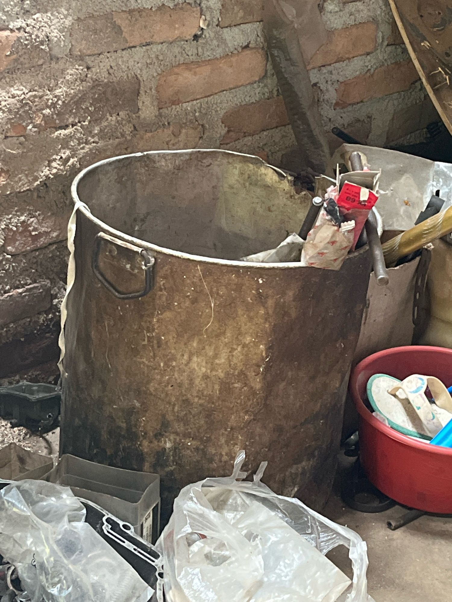 In Kebumen, Indonesia, an interior scene depicts a cluttered storage area. A large, weathered metal bin with two handles dominates the center, filled with various items. To the right, a red plastic basin contains a pair of flip-flops and a blue object, possibly a bottle. Behind the bin, a brick wall forms the backdrop, its mortar rough and uneven. The floor is concrete, strewn with debris including plastic bags filled with unknown contents, and a dark, square object. The overall impression is one of disarray and neglect.