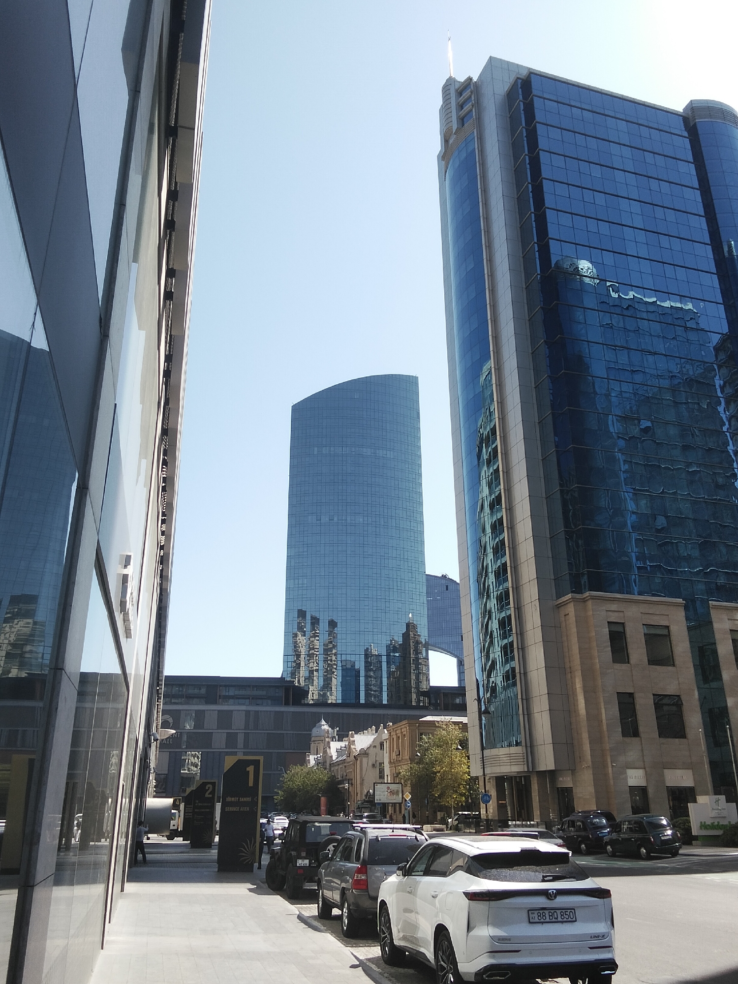 The image shows a street scene in Baku, Azerbaijan, with modern skyscrapers dominating the skyline. On the left, a glass-facade building reflects the sky and surrounding structures. To the right, a tall, blue-glass skyscraper stands prominently, with its reflective surface mirroring the urban environment. Further in the distance, another curved glass skyscraper is visible, and between them, a bridge-like structure connects two buildings. The street level features a sidewalk with several parked cars, including a white SUV in the foreground with a license plate reading "88 BQ 850". Signs indicating "1 XIDMET SAHESI" and "2" are also present on the sidewalk, suggesting a service area. People are sparsely visible, with one individual standing near the sidewalk and another near a parked truck. The overall impression is a bustling urban environment in Baku, characterized by contemporary architecture.