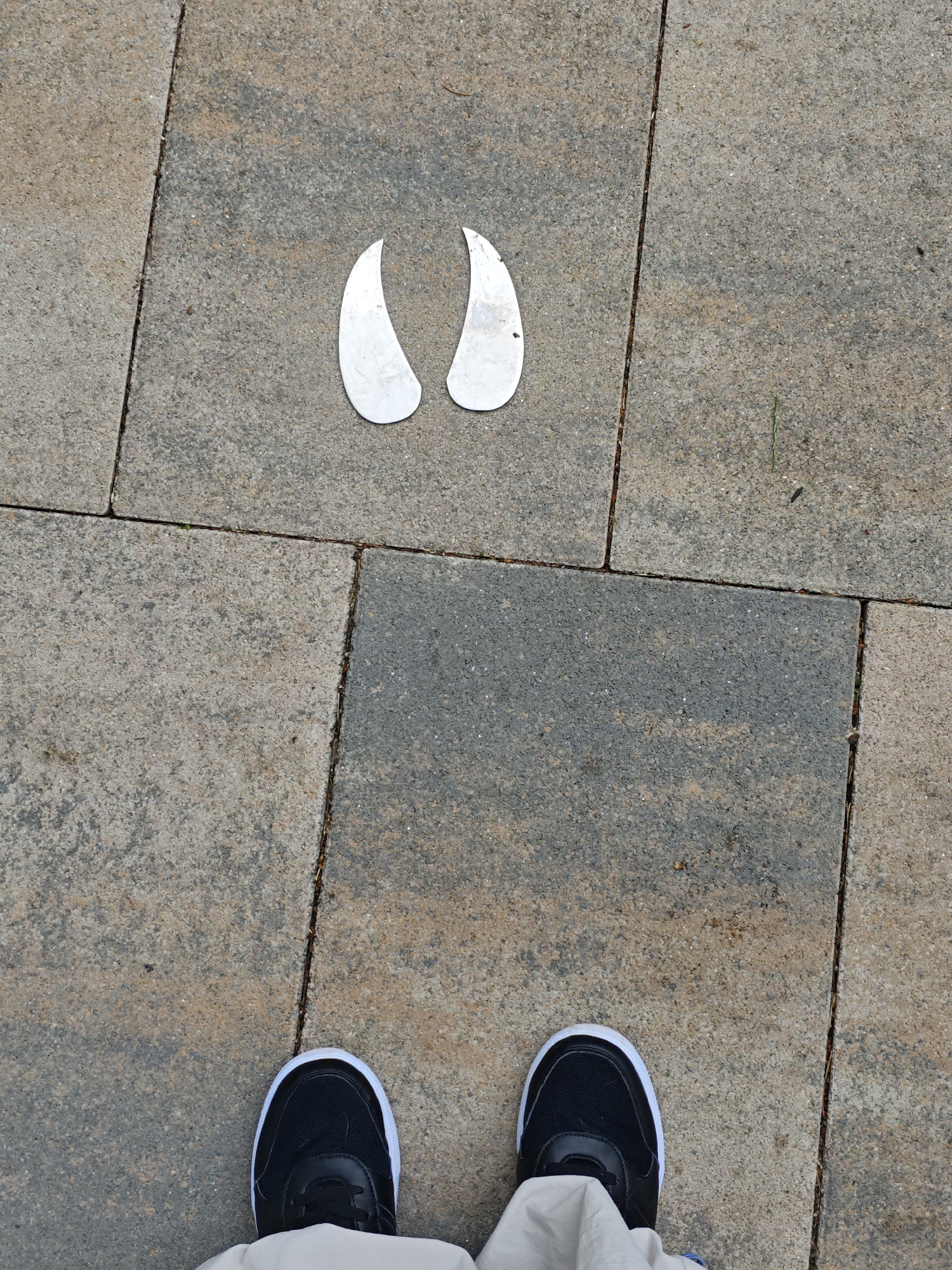 A top-down, eye-level shot shows a pair of black and white sneakers standing on a stone-tiled pathway. The tiles are rectangular and vary in color from light grey to a reddish-brown. Between the tiles, there are narrow gaps filled with dirt and debris. To the upper center of the image, a metallic, two-pronged object resembling a stylized animal hoofprint is embedded in the pathway. The object is silver and has a matte finish. The scene appears to be an outdoor public space, possibly a park or pedestrian area, within the city of Cherkessk, Russia, suggested by the presence of the hoofprint marking which may be part of a local themed trail or educational exhibit.