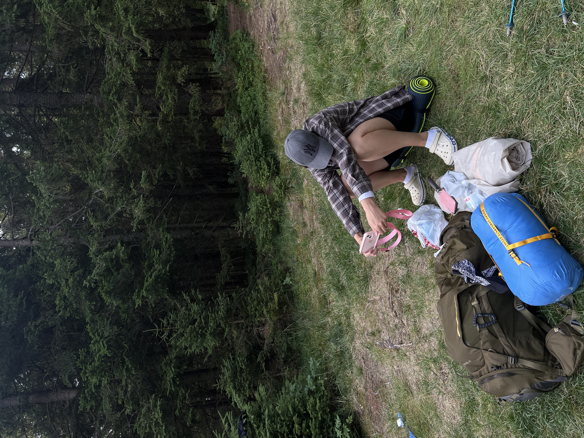 In a forest clearing near Vorokhta, Ukraine, a person wearing a plaid shirt, dark shorts, a baseball cap, and Crocs is crouching. They are holding a pink phone and a pink strap, appearing to be checking or adjusting them. In front of the person, arranged on the grassy ground, are several items typically associated with camping. These include a large olive-green backpack, a rolled-up blue sleeping bag, a rolled-up beige sleeping mat, a rolled-up tent bag, and a black bandana. The surrounding environment is characterized by dense coniferous trees and overgrown grass, indicative of a natural, undeveloped setting. The lighting suggests it is daytime, possibly overcast.