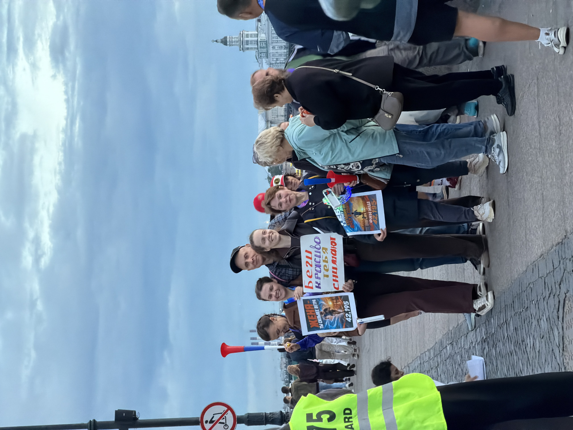 A group of people stand on a cobblestone street near a body of water under a cloudy sky. The individuals are holding signs with text and images. One sign shows a runner and the number "42,195." Another sign has text in purple and white, reading "BEГИ КРАСИВО ТЕБЯ СОЧУВСТВУЮТ." A person in a bright yellow vest with the word "GUARD" is visible on the left. In the background, a building with a tower and smokestacks can be seen. A red and white air horn is held by one of the individuals.