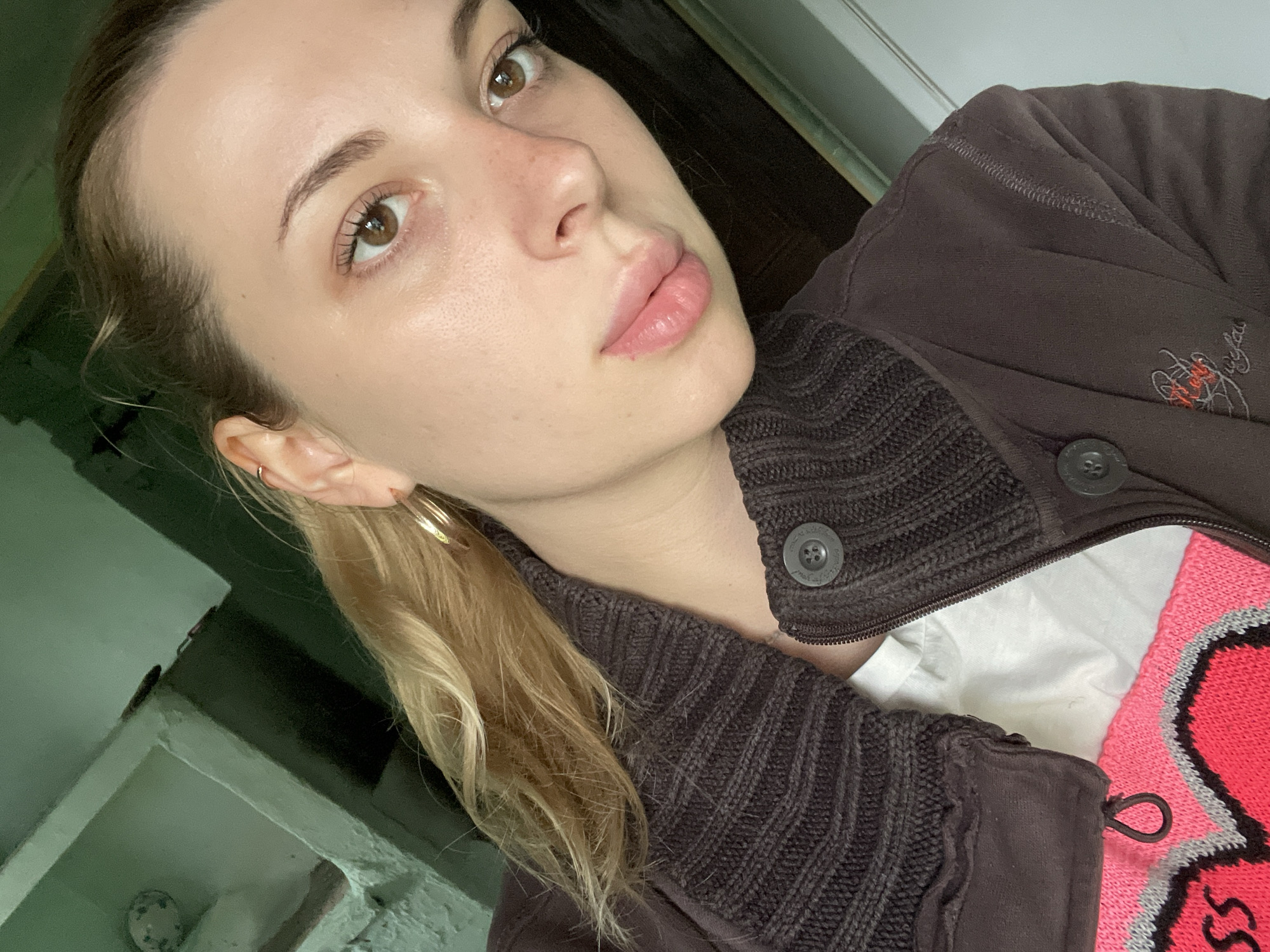 The image is a close-up selfie of a young Caucasian woman with blonde hair pulled back. She is wearing a brown zip-up jacket over a white t-shirt with a bright pink and grey heart design. She has light brown eyes and light skin with some freckles. The background appears to be an indoor setting with green painted walls and shelves, suggesting a modest living space. The overall impression is casual and informal.