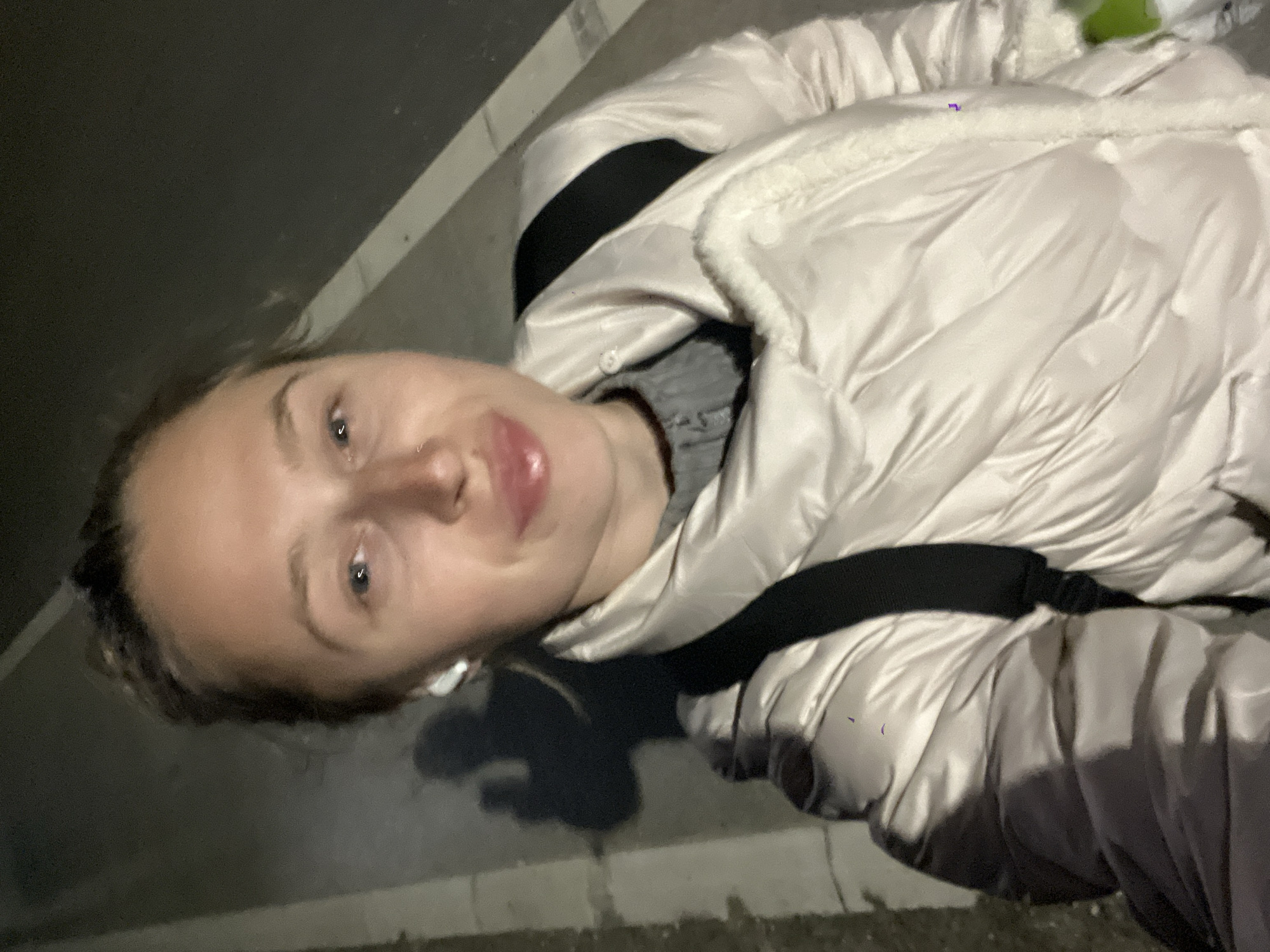 A woman is posing for a selfie outdoors in Boreti, Montenegro. She is wearing a white, padded jacket with a fur-lined hood and a black strap over her shoulder. She has light skin, blue eyes, and her lips are slightly parted. An earbud is visible in her left ear. The background shows a dark asphalt surface with a light-colored curb. The lighting suggests it is nighttime.