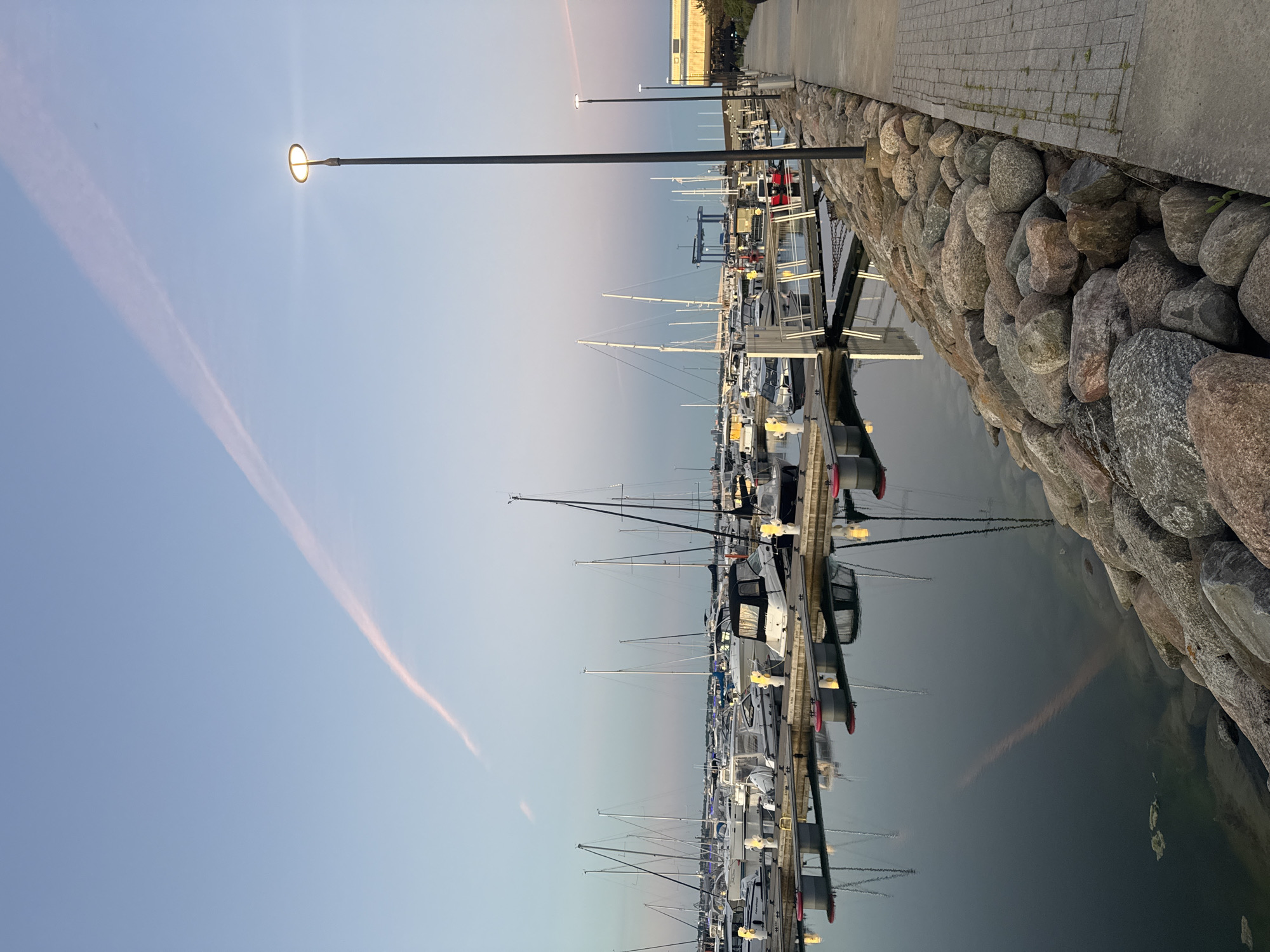 This is a horizontal, eye-level, medium shot of a marina in Tallinn, Estonia, during what appears to be either sunrise or sunset, as indicated by the soft, warm hues in the sky and the gentle illumination of the scene. The sky is a pale blue, with a thin, pinkish contrail streaking across it. In the foreground, a paved walkway runs along the right side of the image, bordered by a rocky embankment. A tall, modern lamppost with an illuminated, ring-shaped head stands prominently on the walkway.
The marina itself is filled with numerous docked sailboats and motorboats, their masts and hulls creating a dense, layered composition. The boats are moored to floating docks, which are clearly visible with red buoys attached. The water in the marina is calm and reflects the boats and the sky, creating a mirror-like effect. A few pieces of floating debris are visible in the water near the rocky embankment.
In the background, beyond the marina, there are hints of urban development with buildings and what appear to be industrial structures, including a large blue crane. The overall impression is one of a peaceful maritime setting within an urban environment. No people are clearly visible in the image.