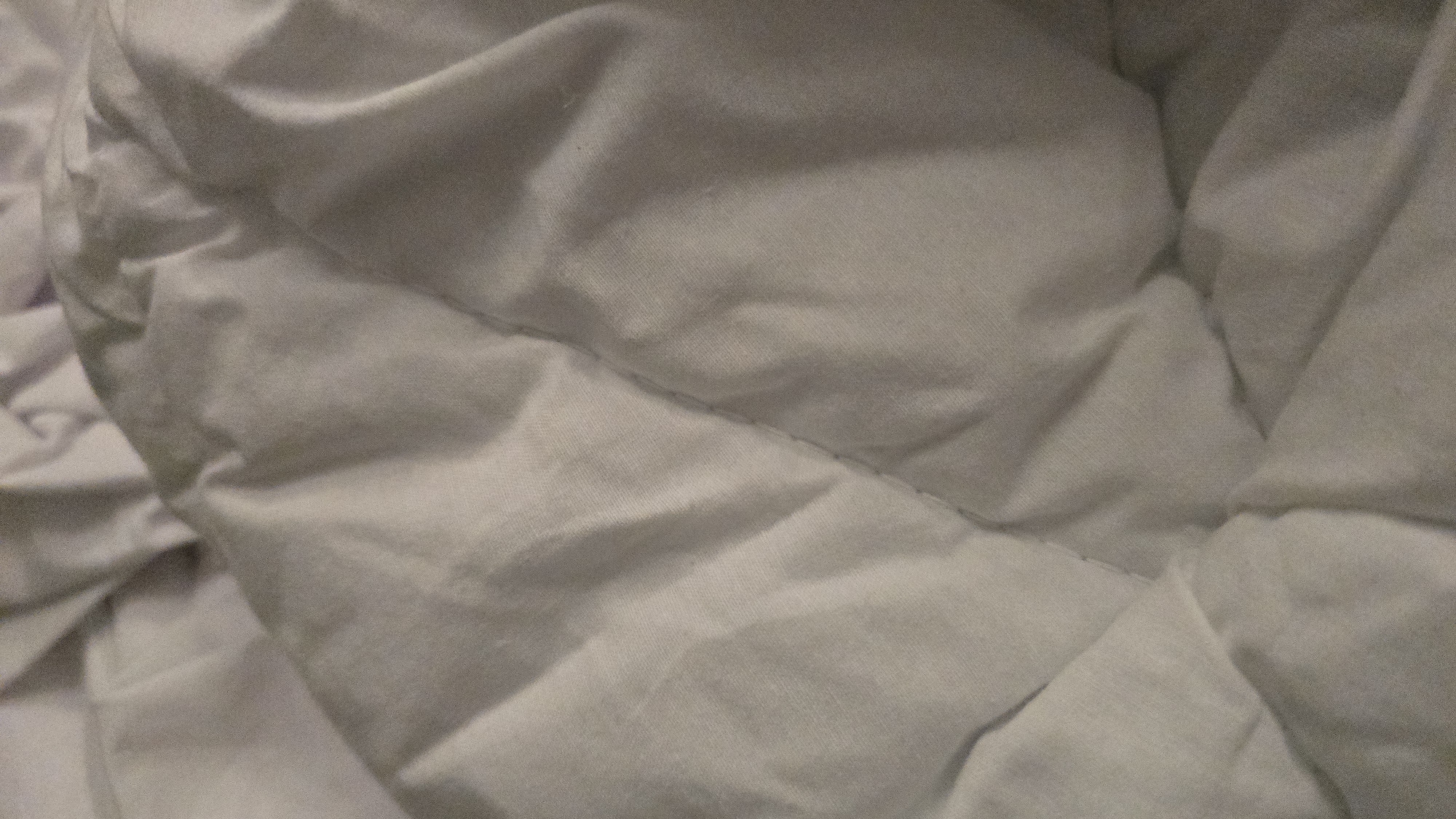 The image is a close-up, low-angle shot of a white comforter or duvet. The fabric is rumpled and creased, creating a textured surface with shadows and highlights. A visible stitch line runs diagonally across the frame from the upper left to the lower right, dividing the fabric into two sections with different folding patterns. The overall impression is one of soft, folded fabric. The location of this scene within the city of Batumi, Georgia, cannot be determined from the provided image.