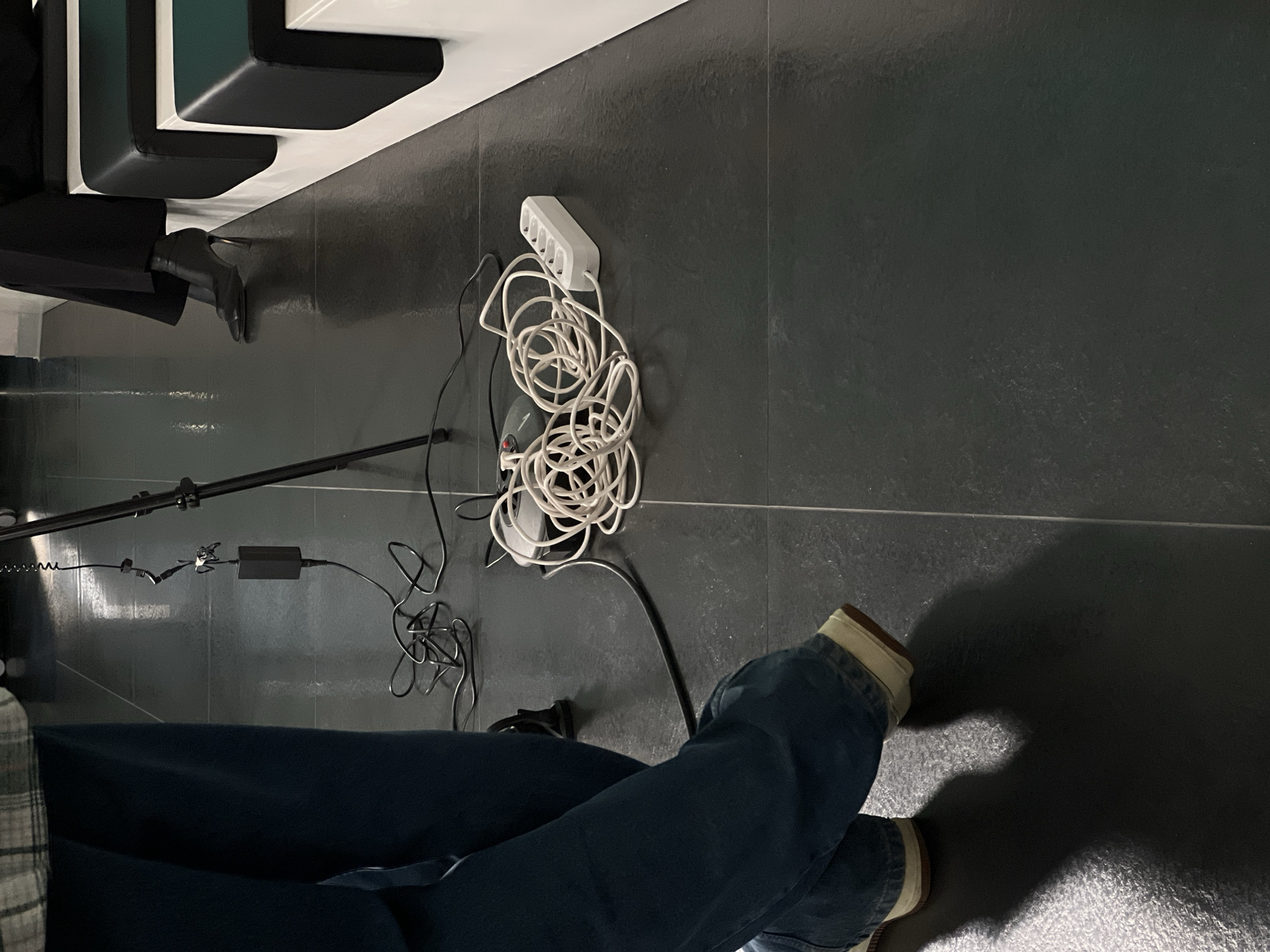 The image depicts an indoor setting with a dark grey tiled floor, featuring two individuals and electronic equipment. No specific architectural or contextual elements are visible to identify a precise location within the city of Moscow, Russia.
In the left foreground, a person is shown from the mid-thigh down, wearing blue denim jeans and light-colored sneakers with tan soles; their legs are crossed at the ankles. A portion of a plaid garment is visible on their upper body. Centrally, a black camera tripod stands, with a black power adapter connected by a coiled black cable hanging from it. Beneath the tripod, a tangle of black electrical cables, a coiled white extension cord, a grey electronic device, and a white five-outlet power strip lie on the floor. To the right, the lower legs of a second individual are visible, seated on a modular white bench with black and dark green upholstered cushions. This person wears black wide-leg trousers and black high-heeled ankle boots with metallic heels. A white wall forms the background on the right side of the frame.