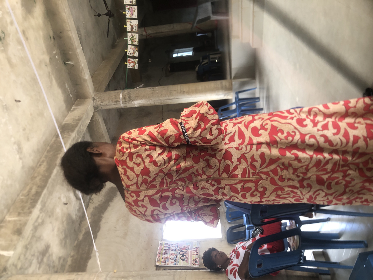 A person stands on the right side of the frame, seen from the back and slightly to the left, wearing a long-sleeved dress patterned with red and light beige/gold intricate designs. Their dark hair is pulled back. In the mid-ground, on the left, another person is seated on a blue plastic chair, wearing a white top with red patterns and red trousers. This seated person has short, dark, curly hair and appears to be looking towards the right; a small black object, possibly a microphone, is visible near their lap. Several other blue plastic chairs are stacked or arranged around the room.
The setting is an indoor space with bare concrete walls and ceiling, featuring visible concrete beams and supports. A dark ceiling fan is mounted in the upper left corner. A white string or wire stretches across the upper portion of the room, from left to right, with small rectangular cards or pictures clipped to it; one card appears to show the letter 'N'. A window in the mid-right background lets in bright daylight, and a staircase is visible ascending in the far upper right. On a wall in the lower left, there is a poster or collage of small images, possibly portraits. The overall impression is that of a functional community hall or classroom, possibly for a gathering or meeting.
Location context: Unknown, Unknown