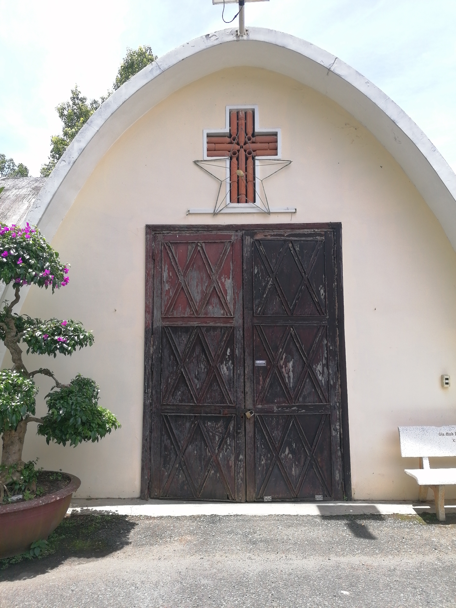The image shows a weathered, dark wooden double door with diamond-shaped patterns on a pale yellow, arched building, likely a church or chapel. Above the door, a cross made of terracotta-colored tubes is mounted on the wall, partially enclosed by a star-shaped metal frame. A small bonsai tree with vibrant purple flowers sits in a large reddish-brown pot to the left of the door. To the right, a white stone bench with an inscription is visible against the building's wall. The ground in front of the building is asphalt, with a patch of gravelly dirt. The scene is illuminated by natural daylight, with shadows cast on the ground. The building appears to be located in a relatively quiet, outdoor setting.