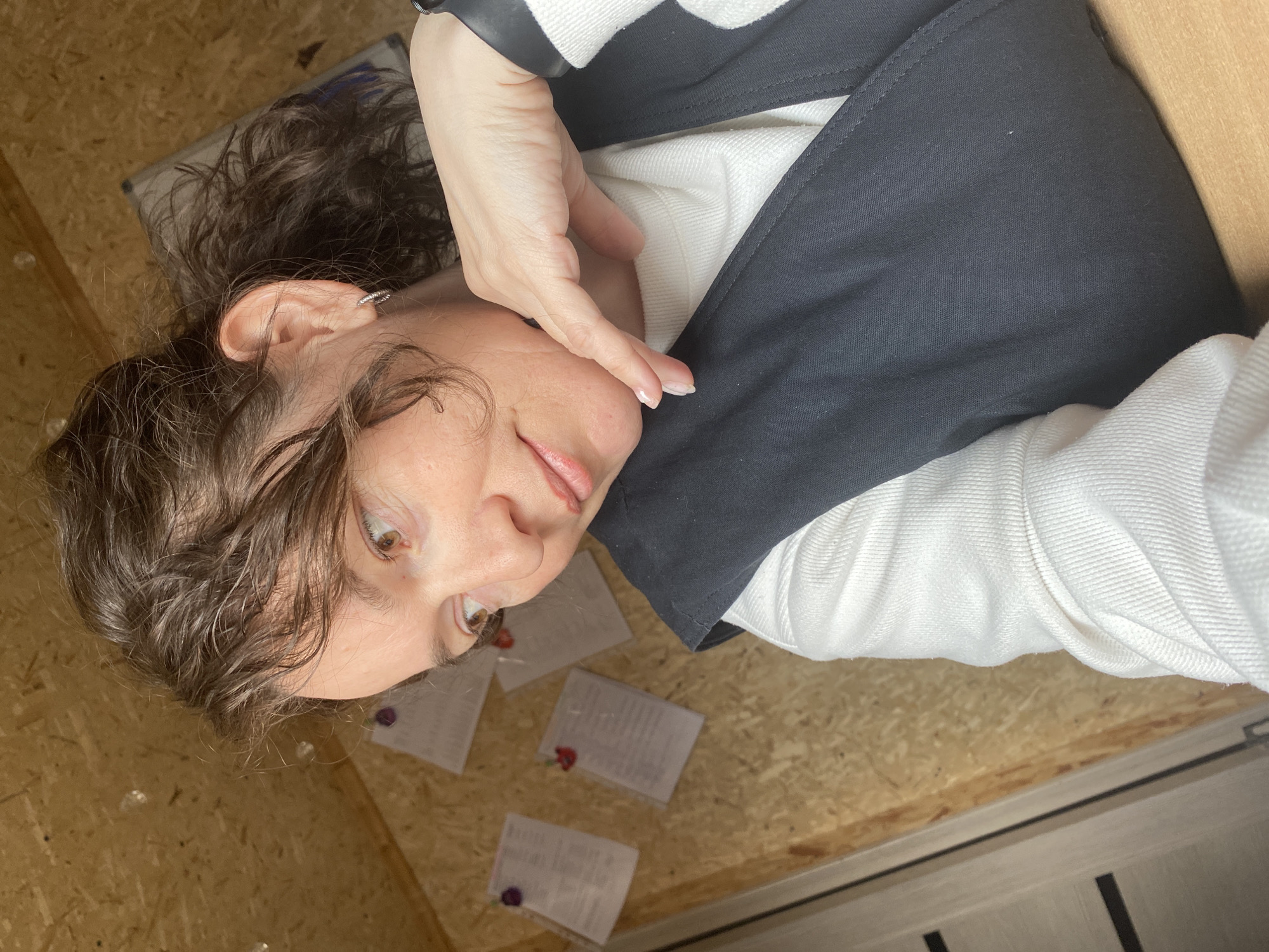 A woman lies on a surface that appears to be plywood, with several small documents scattered nearby. She is wearing a white sweater with a black vest or jacket. Her hand touches her face. The location is unknown, and the presence of a subject in Ol'khovskoye, Russia cannot be confirmed.