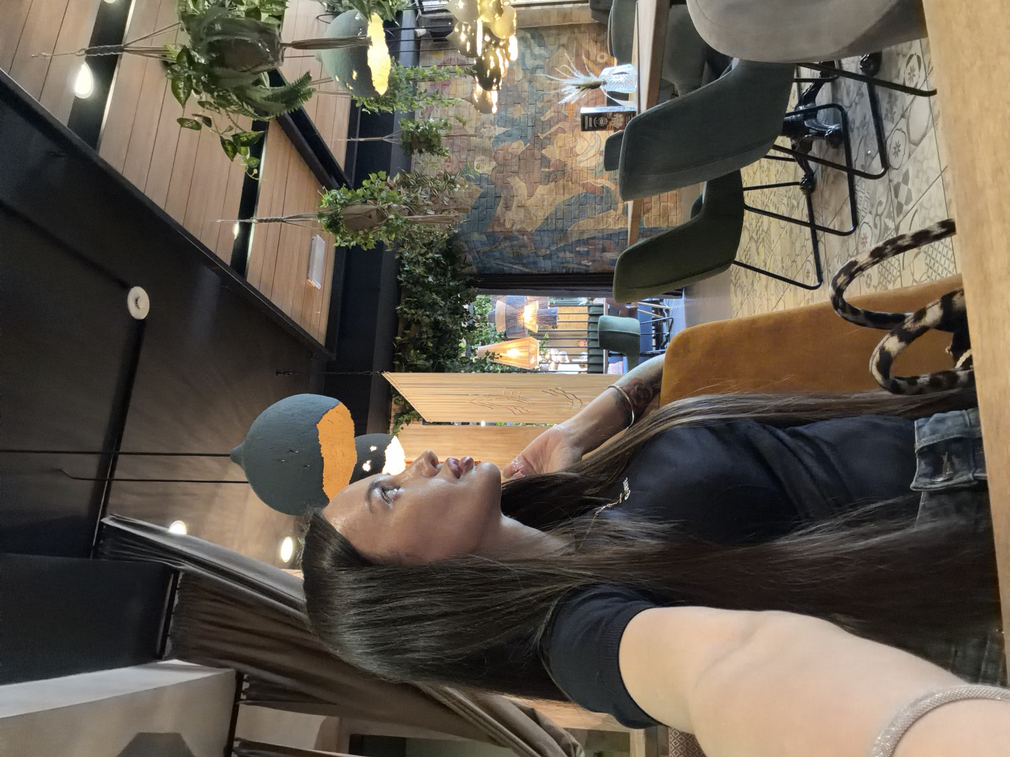 This image, rotated 90 degrees counter-clockwise, depicts a woman looking upwards within what appears to be a stylish indoor setting, likely a cafe or restaurant.
The primary subject is a woman positioned on the right side of the frame. She has long, dark brown hair flowing down her back and over her shoulder, and fair skin. She is wearing a black t-shirt with faint white text on the chest, which is not clearly legible. Her gaze is directed upwards and slightly to her right, with a calm, contemplative expression. Her right arm is bent, with her hand near her face, and her left wrist features a silver-toned bracelet. She appears to be posing for a photo or taking a selfie.
The background reveals a sophisticated interior design. The ceiling features dark wooden beams and lighter wooden panels, from which various decorative elements hang. Several unique pendant lights, dark grey or black spheres with orange inner surfaces, are visible near the woman. Further back, more elaborate chandeliers with multiple warm-toned bulbs illuminate the space. Hanging plants with green foliage are suspended throughout the area, adding a natural touch.
One wall in the background is adorned with a large, colorful mural or tiled artwork depicting abstract shapes and possibly figures in muted tones of blue, beige, and terracotta. The floor is covered with patterned tiles in shades of grey and white. Furniture includes several modern chairs with dark grey upholstered seats and black metal frames, and a chair with a warm brown/orange upholstered seat. A leopard-print patterned strap or handle is visible near the brown chair. The overall lighting is warm, suggesting an evening or atmospheric indoor setting.