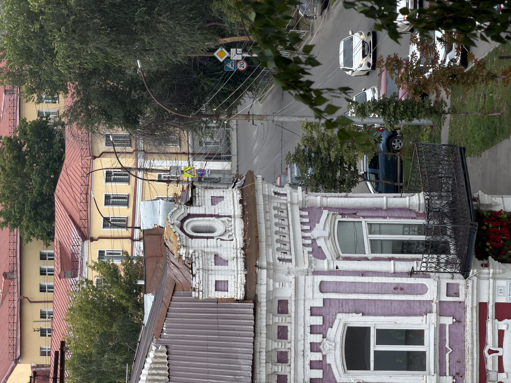 The image shows a street scene in Saratov, Russia. In the foreground, a ornate purple and white building with a corrugated metal roof is visible. A wrought-iron balcony extends from the building. The street below is lined with trees and utility poles with numerous wires. Several cars are parked or driving on the street, including a white sedan, a dark blue SUV, and a red car. In the background, there are yellow buildings with red roofs. Traffic signs are also visible, including a pedestrian crossing sign, a prohibition sign, and a speed limit sign indicating 20.