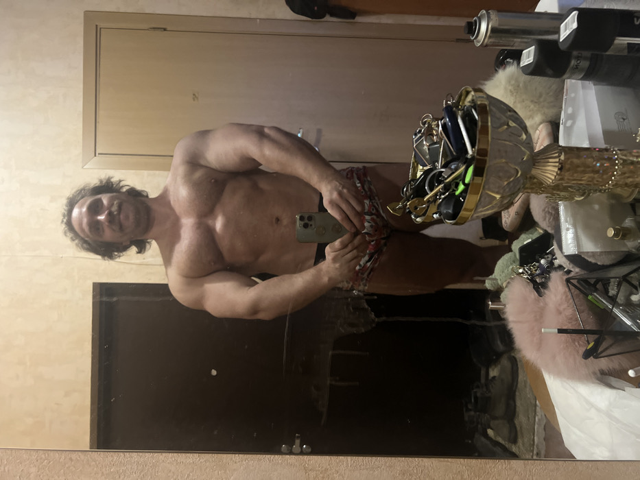 A muscular man with long, wavy hair and a slight mustache poses shirtless in a mirror, holding a smartphone. He wears patterned shorts and has a visible abdomen. The mirror reflects a messy background with various items, including a gold decorative bowl filled with keys and other objects, spray cans, and possibly clothing. The room has a neutral color palette.