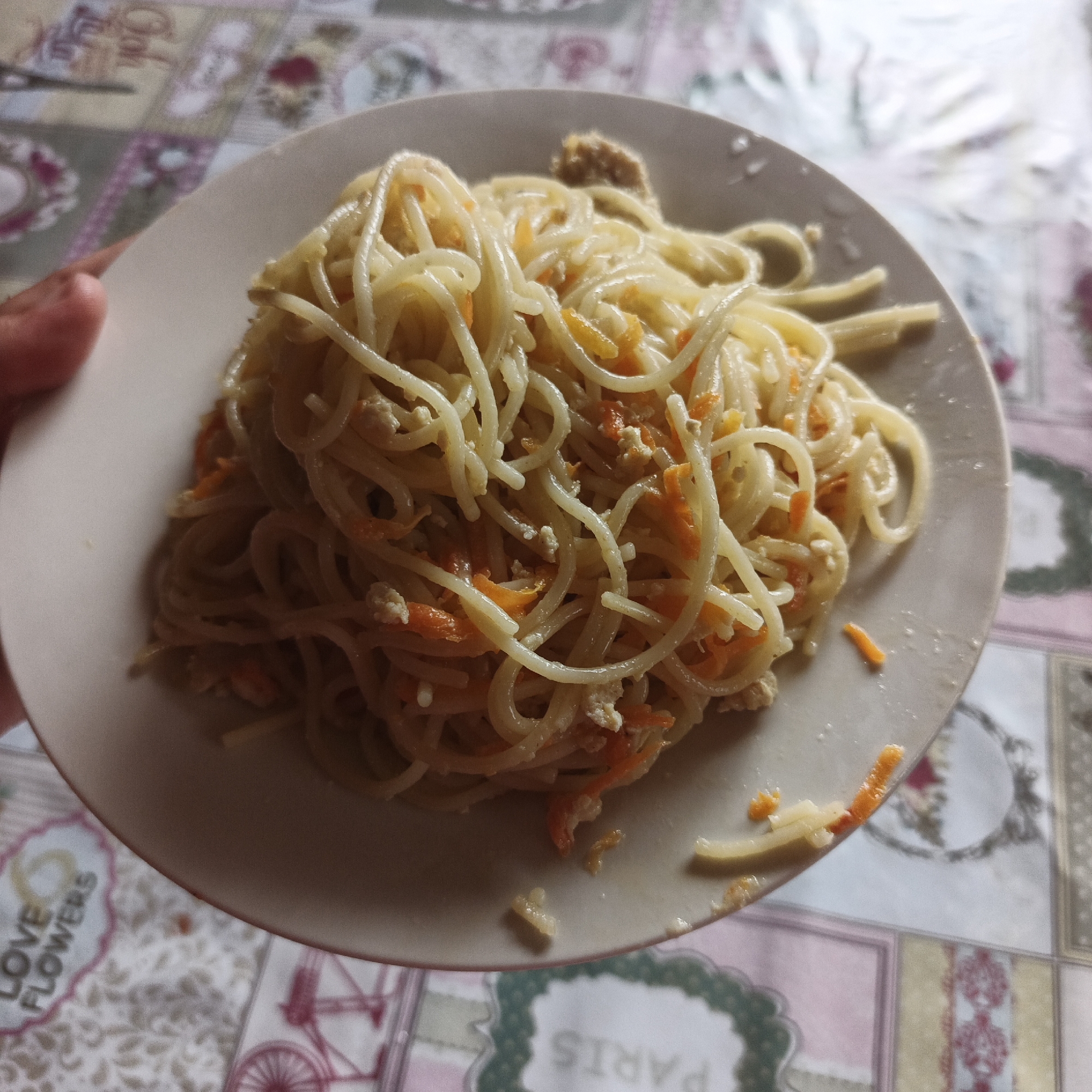 A close-up overhead shot shows a plate of spaghetti topped with shredded carrots and crumbled chicken or tofu. The plate is on a colorful, patterned tablecloth. A hand is visible on the left edge of the frame, holding the plate. The spaghetti is light in color, with thin strands coated in a sauce. The carrots are bright orange, and the protein appears white and crumbly. The tablecloth has a floral pattern with pink, white, and brown colors. The scene is illuminated by natural light, casting soft shadows. The image does not provide any information about the location in the city of Podlubovo, Russia.