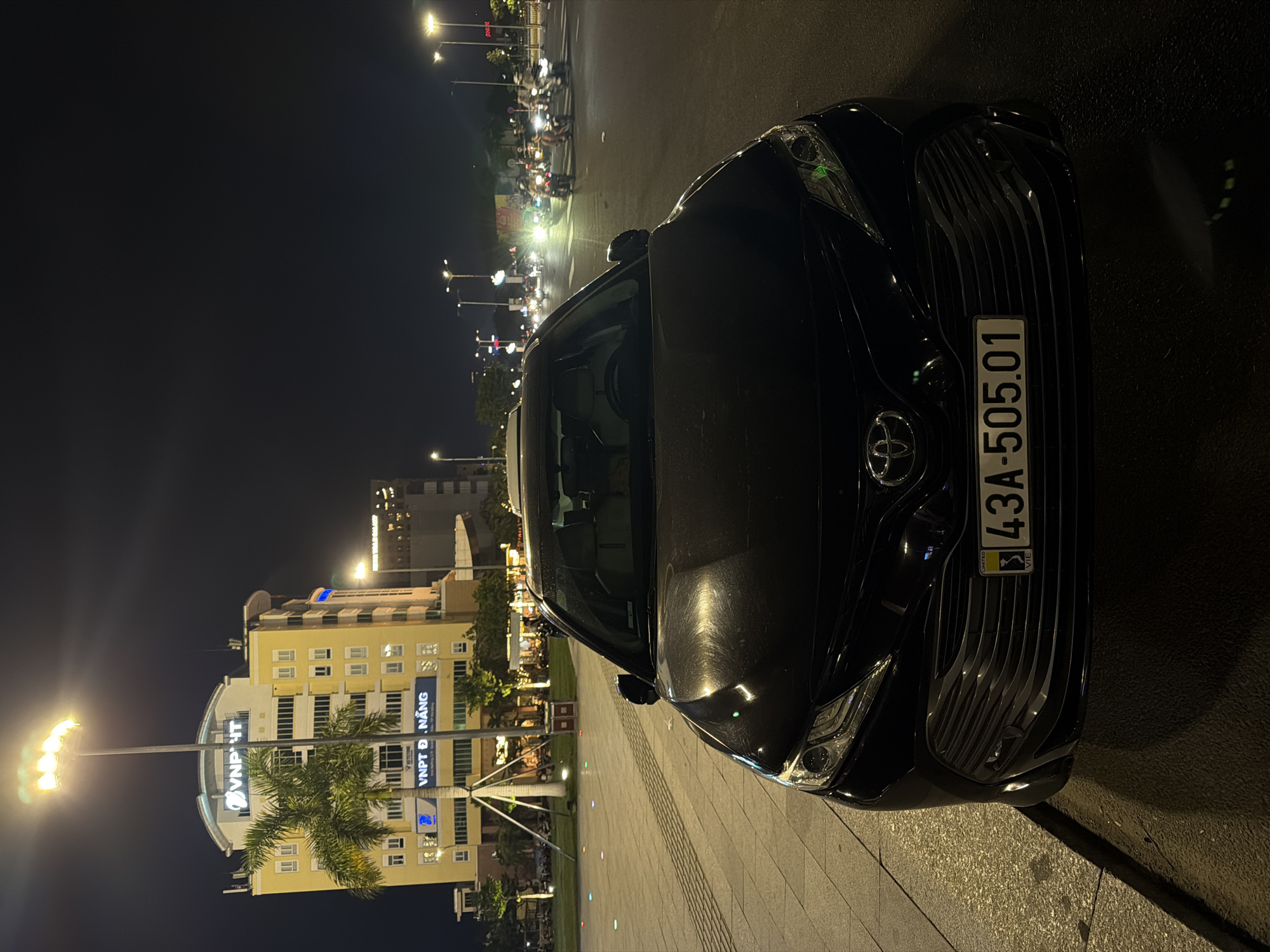 A black Toyota sedan is parked on a street at night in Da Nang, Vietnam. The car is facing forward, and the hood and front grille are prominently featured. The license plate reads "43A-505.01". In the background, a yellow building with the "VNPT" logo is visible, along with palm trees and streetlights. A street with multiple motorcycles is also visible in the background, indicating it's an urban environment. The scene is illuminated by artificial lights, suggesting it is nighttime.