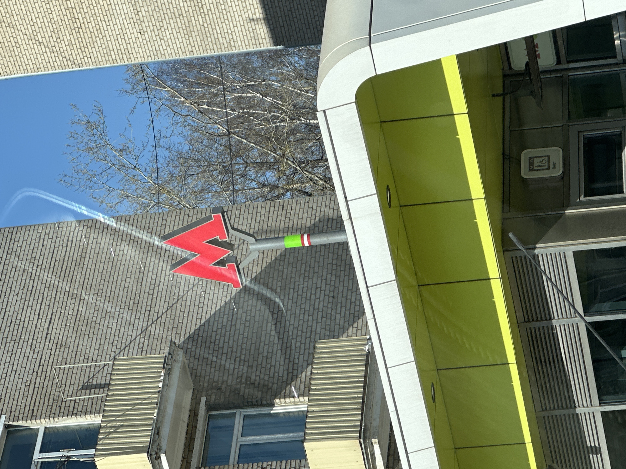The image shows a metro sign with a red letter "M" in Moscow, Russia. The sign is attached to a gray brick building, with a leafless tree visible in the background. A modern, green and white building is in the foreground. The sky is blue.