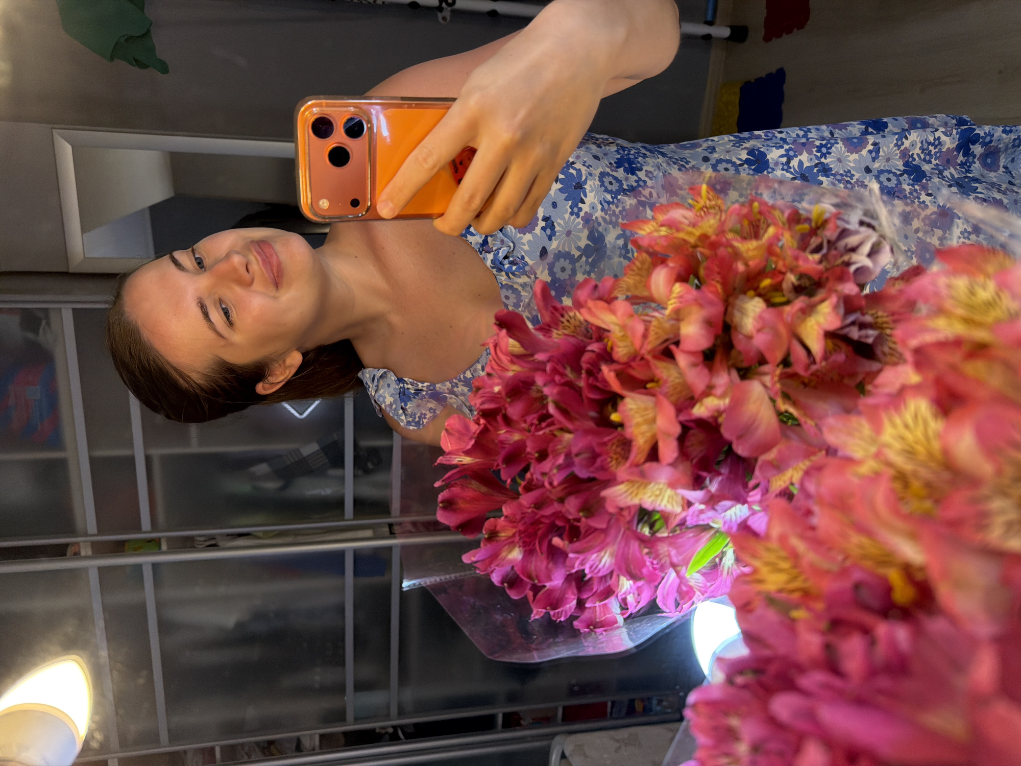 A young woman is captured in a mirror selfie, holding a large, vibrant bouquet of flowers. She is smiling gently, looking directly into the phone's camera. Her dark hair is pulled back, and she is wearing a blue dress adorned with a white floral pattern.
In her right hand, she holds a smartphone encased in a bright orange cover, its triple camera lenses clearly visible on the back. The bouquet she holds is prominent, dominated by numerous pink and red alstroemeria (Peruvian lilies) with distinct yellow centers and dark streaking on their petals. The flowers are wrapped in a clear plastic film.
The setting appears to be an indoor space, reflected in what seems to be a large mirror. Behind the woman and to her left, there are shelves or cabinet structures, and a light fixture with a warm glow is visible in the lower left corner. The overall lighting suggests an indoor environment. There is no visible text in the image. The scene conveys a cheerful and pleasant mood, likely capturing a moment of personal happiness or appreciation for the flowers.