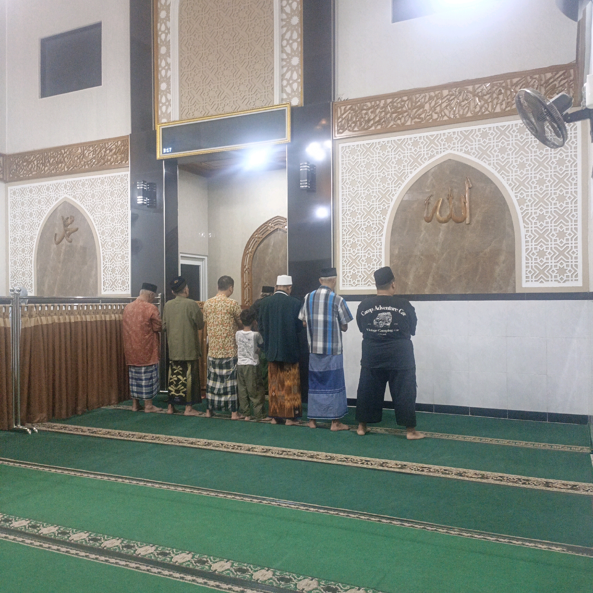 The image shows the interior of a mosque in Salatiga, Indonesia, with a group of people standing in prayer on a green carpeted floor. The mosque features traditional Islamic architectural elements, including ornate patterned walls, arched niches with Arabic calligraphy, and decorative wooden lattices. A fan is mounted on the wall, and a digital display is visible on the wall. The people are dressed in traditional Islamic attire, including sarongs and caps. They are facing towards the qibla, the direction of prayer. The scene suggests a moment of religious observance and community within the mosque.
