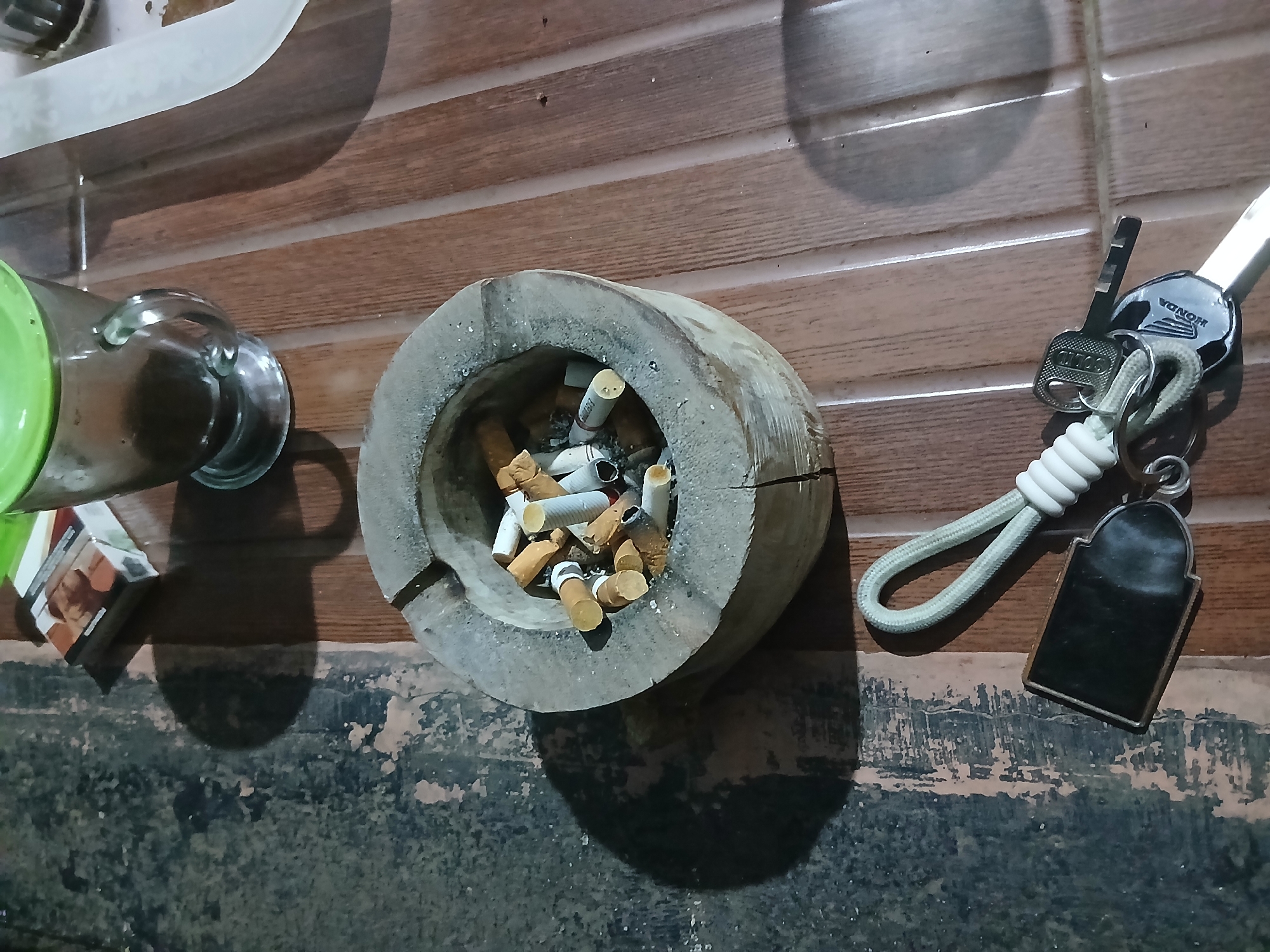 The image shows a close-up, overhead view of a wooden ashtray filled with cigarette butts, a pack of cigarettes, a glass mug with a green lid, and a set of keys. The scene appears to be outdoors, possibly on a porch or patio, with a textured, dark surface on the left and a wooden planked surface on the right. The lighting suggests it is nighttime or a dimly lit interior. The ashtray is centrally located, overflowing with discarded cigarette butts, some of which are partially burned. To the upper left of the ashtray is a pack of cigarettes. To the upper right, a glass mug with a green lid contains a dark liquid, likely coffee or tea. In the lower portion of the image, a set of keys is visible, attached to a white lanyard with a silver keychain. One of the keys has the Honda logo. The environment is cluttered but orderly. There are no people visible in the image. The overall impression is of a casual, possibly late-night or early-morning setting.