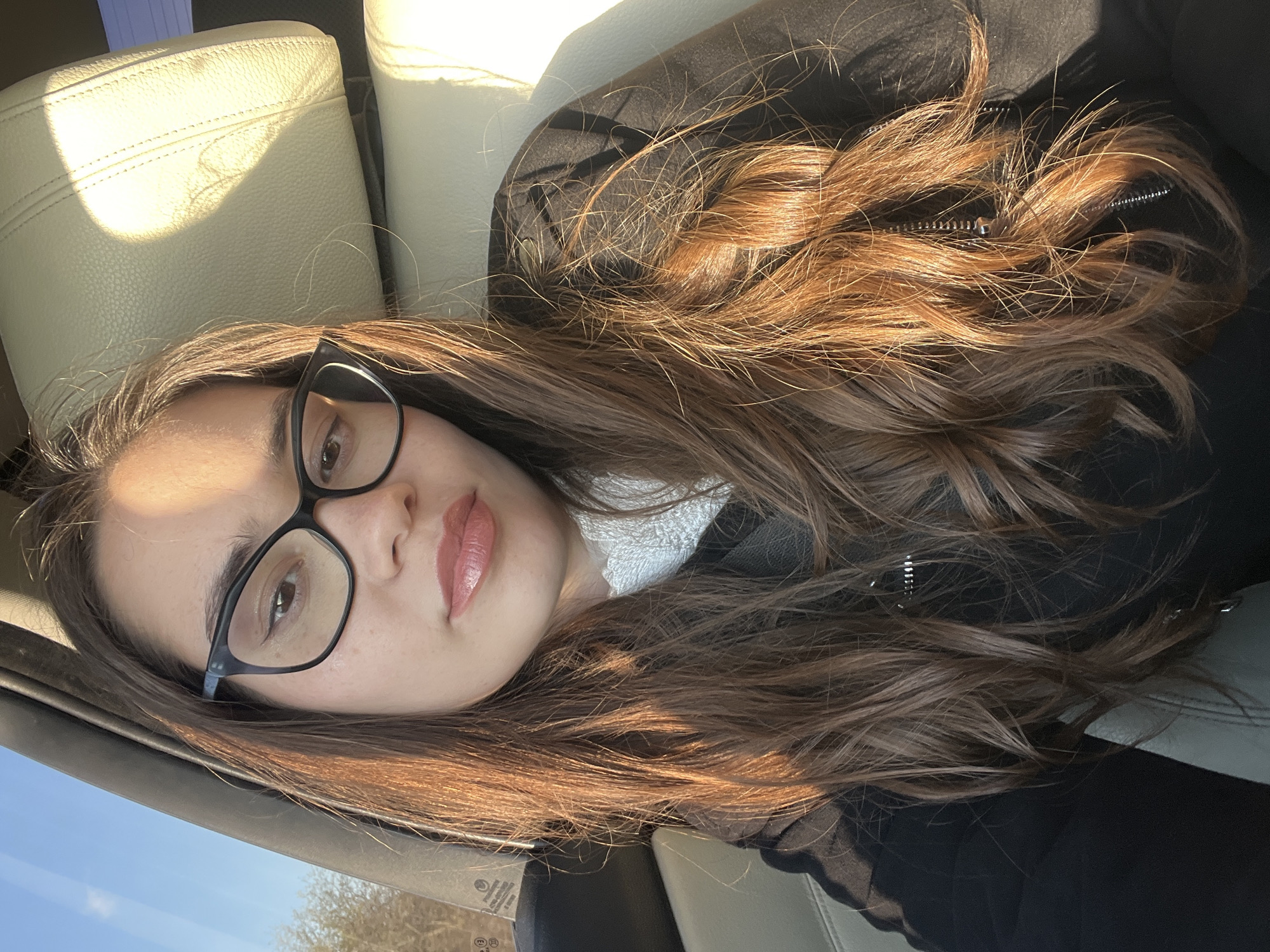 A woman with long brown hair and glasses lies reclined in a car seat. She is wearing a black jacket and a white shirt. The interior of the car is beige leather. Outside the car window, trees and a blue sky are visible. The location is implied, but not visually confirmed, to be Rogachevo, Bulgaria.