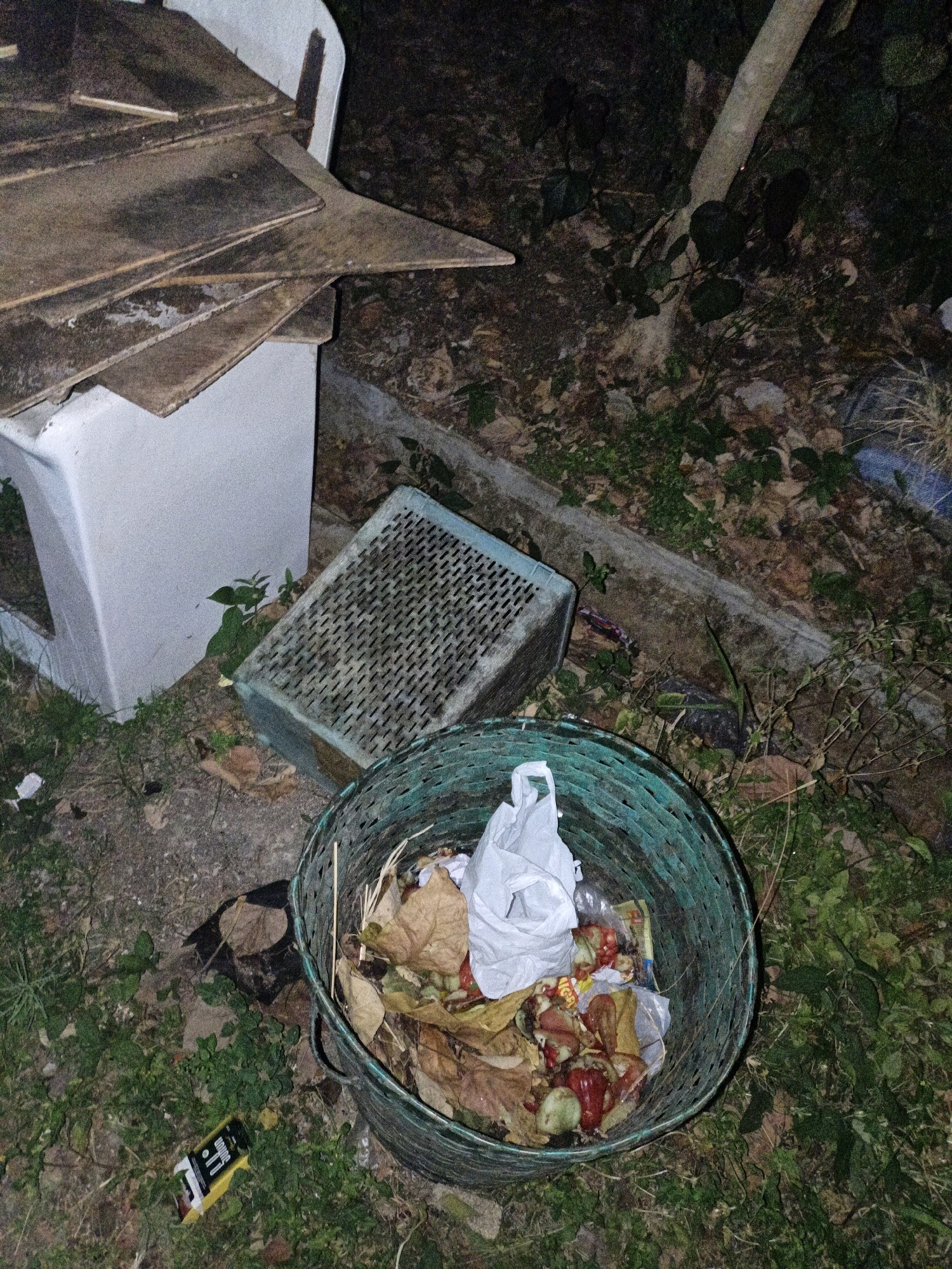 The image depicts an outdoor scene at night, likely in Cikal, Indonesia. In the foreground, a green woven basket is filled with organic waste, including fruit peels, vegetable scraps, and dried leaves. A white plastic bag is also partially visible within the basket. To the left of the basket, a blue-green plastic crate sits on the ground. Behind the crate, a white concrete structure, possibly part of a building or a large planter, is visible. Wooden planks are stacked on top of this structure. The ground is covered with dirt, leaves, and vegetation. A fallen tree trunk or large branch is present in the background, with foliage surrounding it. A yellow wrapper with some text is lying on the ground near the basket. The overall impression is one of an unkempt outdoor area with accumulated refuse.