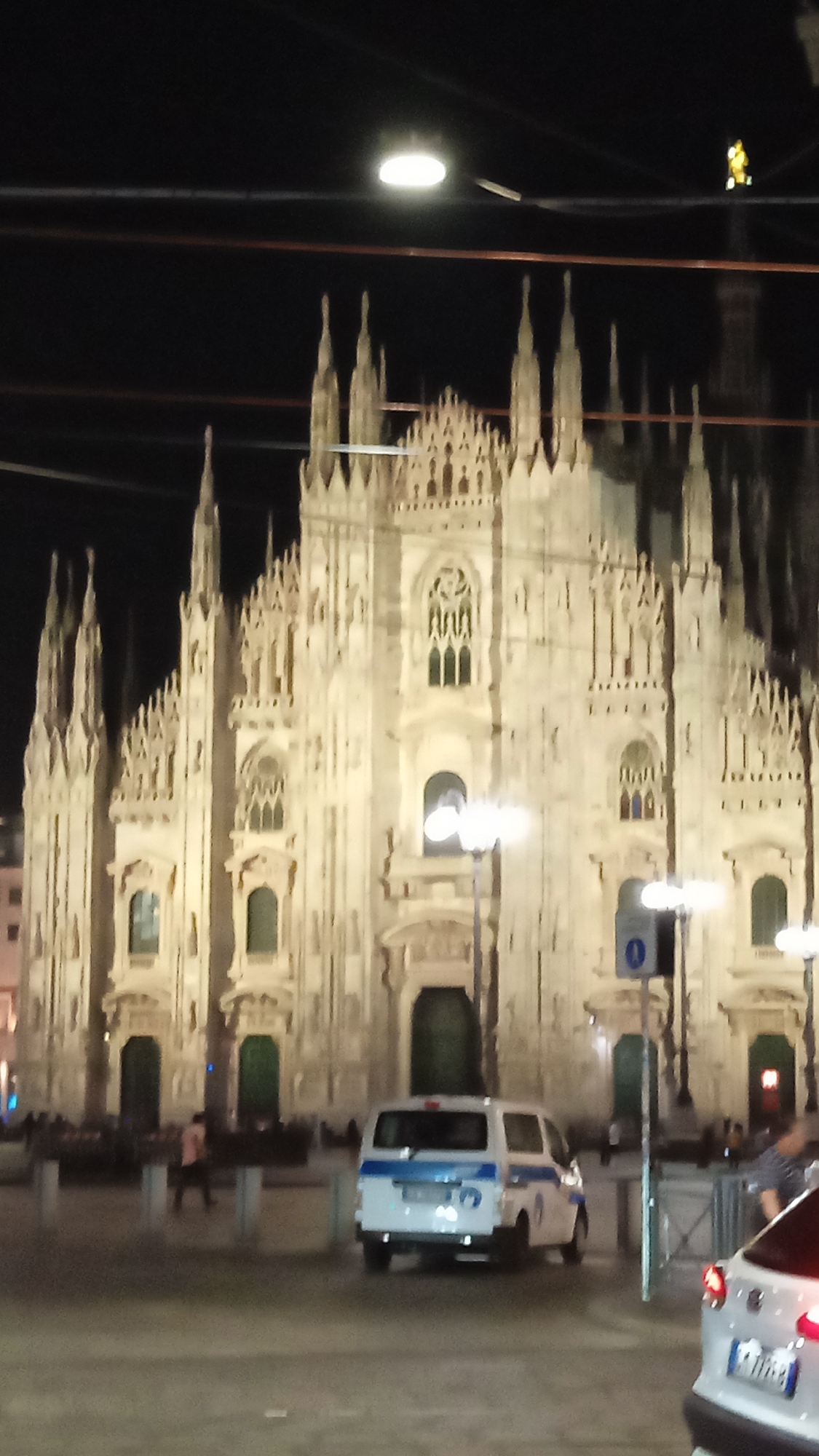 The image shows a night view of the Milan Cathedral (Duomo di Milano) in Milan, Italy. The cathedral's facade is illuminated, with its intricate Gothic architecture prominently visible. In the foreground, a white van with blue stripes, possibly a police vehicle, is parked on the street. Several streetlights illuminate the square. A few people are visible in the background, some walking and others gathered. The overall scene suggests a public space in Milan at night.