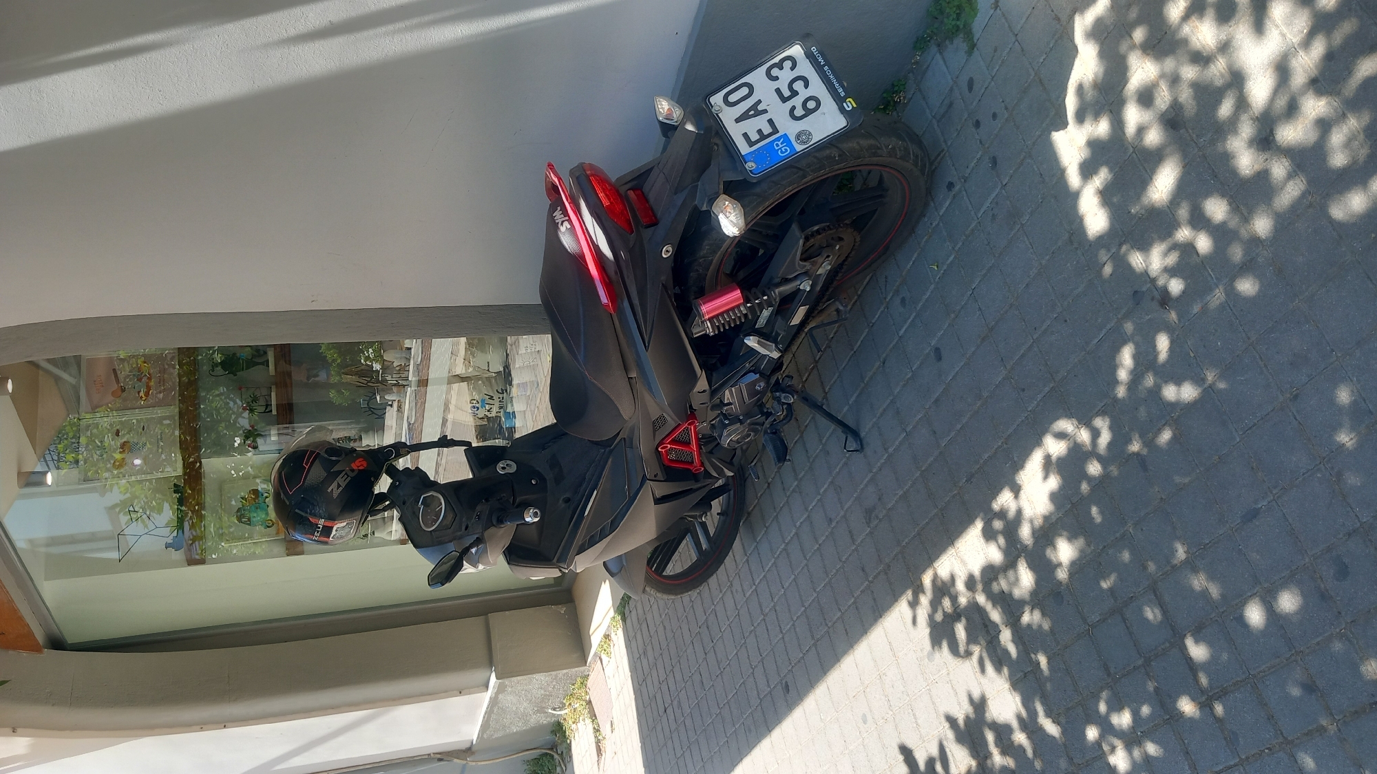 A black SYM scooter is parked on a grey paved sidewalk in front of a building with a large glass window. A black and red helmet rests on the handlebars of the scooter. The scooter has a red accent on the rear suspension and a red strip on the rear fender. A license plate, EAO 653 GR, is attached to the rear of the scooter. The license plate also shows the text "SEDMIKos MOTO". The sidewalk is partially covered in shadow cast by trees. The building appears to be a shop, with displays visible through the window. The scene is set in the city of Kos, Greece.