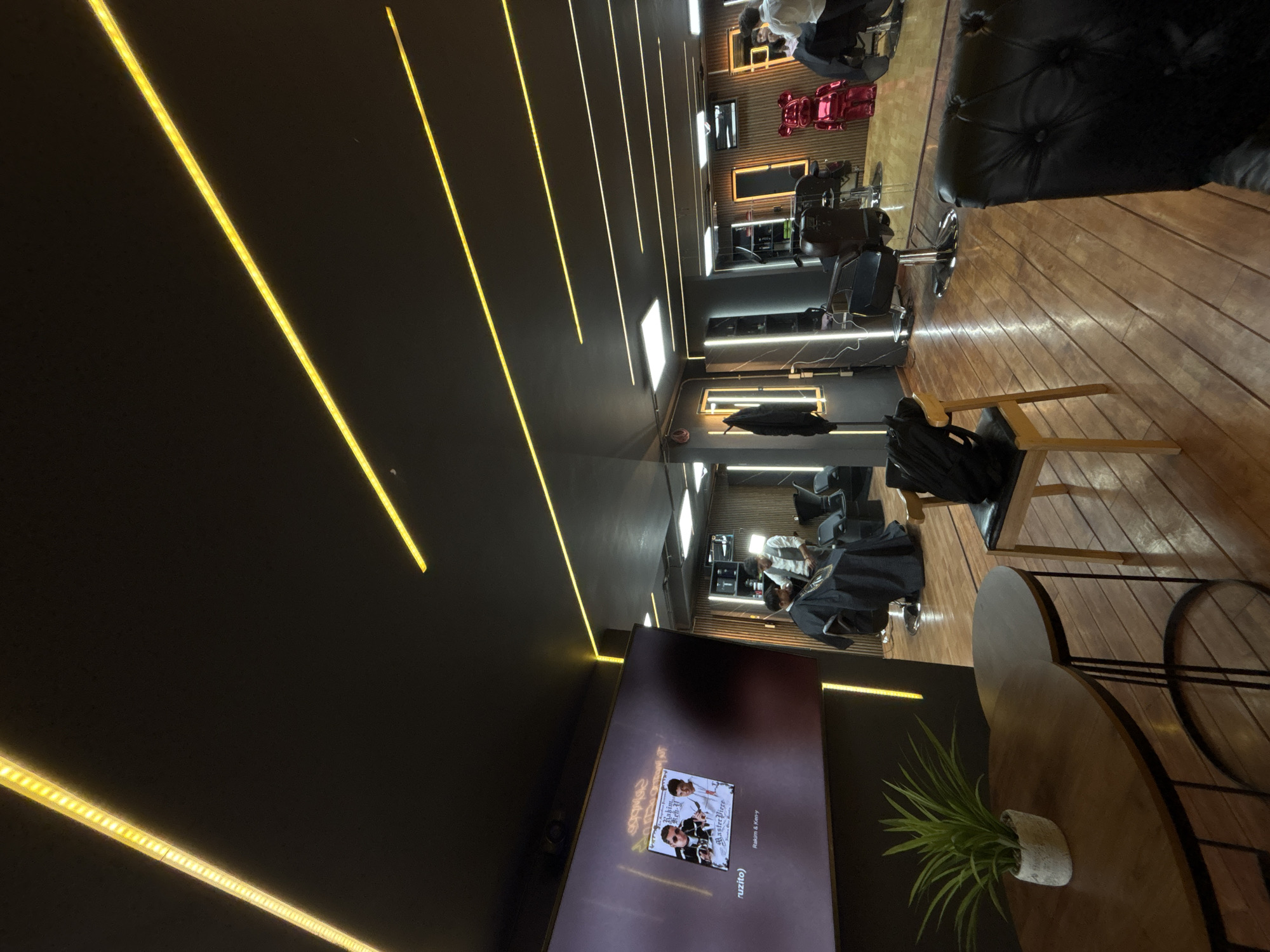 The image captures the interior of a barbershop in Puebla, Mexico. The location has a modern design with dark grey walls and ceiling, illuminated by long, linear LED lights that run along the ceiling. The floor is wood. In the foreground, there is a television mounted on the wall displaying album art. A round wooden side table with a plant sits in front of the TV. A chair is visible beside the table. In the barbershop, chairs and mirrors line the walls. People are seated in barber chairs, some with capes on, implying haircuts in progress. The overall impression is a modern and stylish interior, with a focus on grooming services.