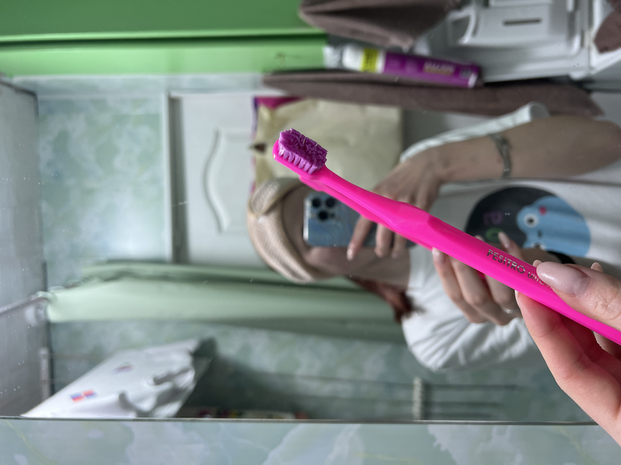 A person is reflected in a mirror while holding a pink and purple toothbrush. The toothbrush is labeled "PESITRO UKIC". The person is holding a smartphone, potentially taking the photo. A white shirt is visible on the person. The surrounding environment includes a medicine cabinet, toiletries, and a green-framed mirror. The image was taken in a bathroom. It is not possible to verify the location is Chaikovsky, Russia.